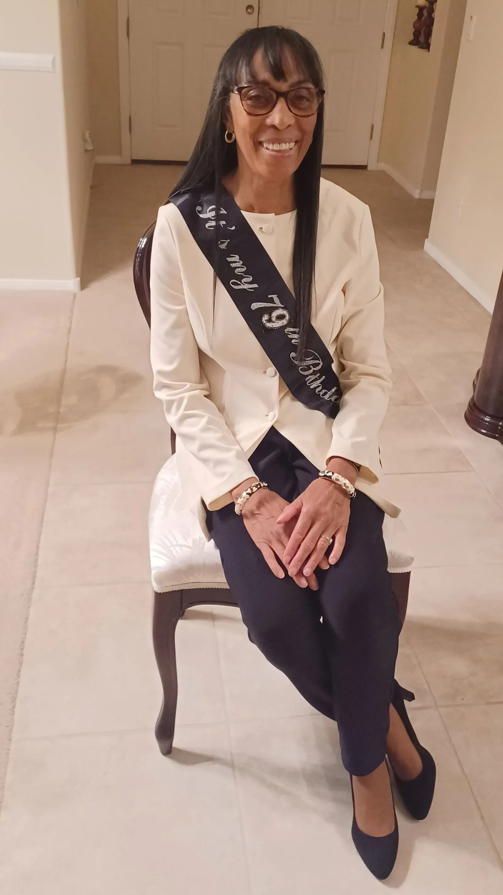 امرأة مسنّة تجلس على كرسي، ترتدي سترة بيضاء وسروالاً داكنًا، تحمل شارة تعلن عن عيد ميلادها السابع والسبعين.