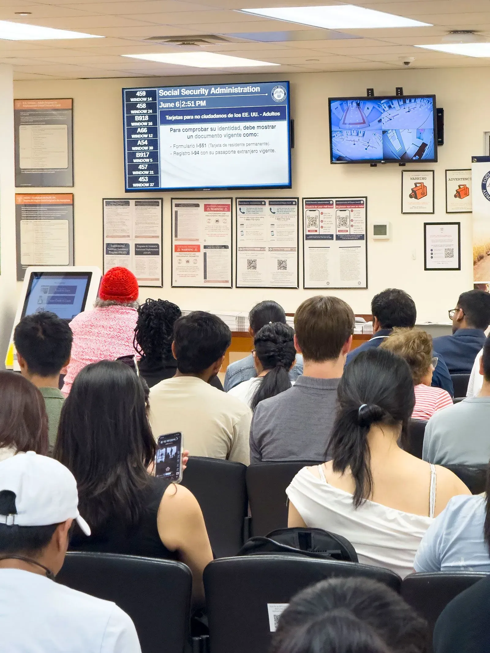 صورة لقاعة انتظار إدارة الضمان الاجتماعي، حيث يجلس العديد من الأشخاص في صفوف، مع شاشة تعرض معلومات الخدمة.