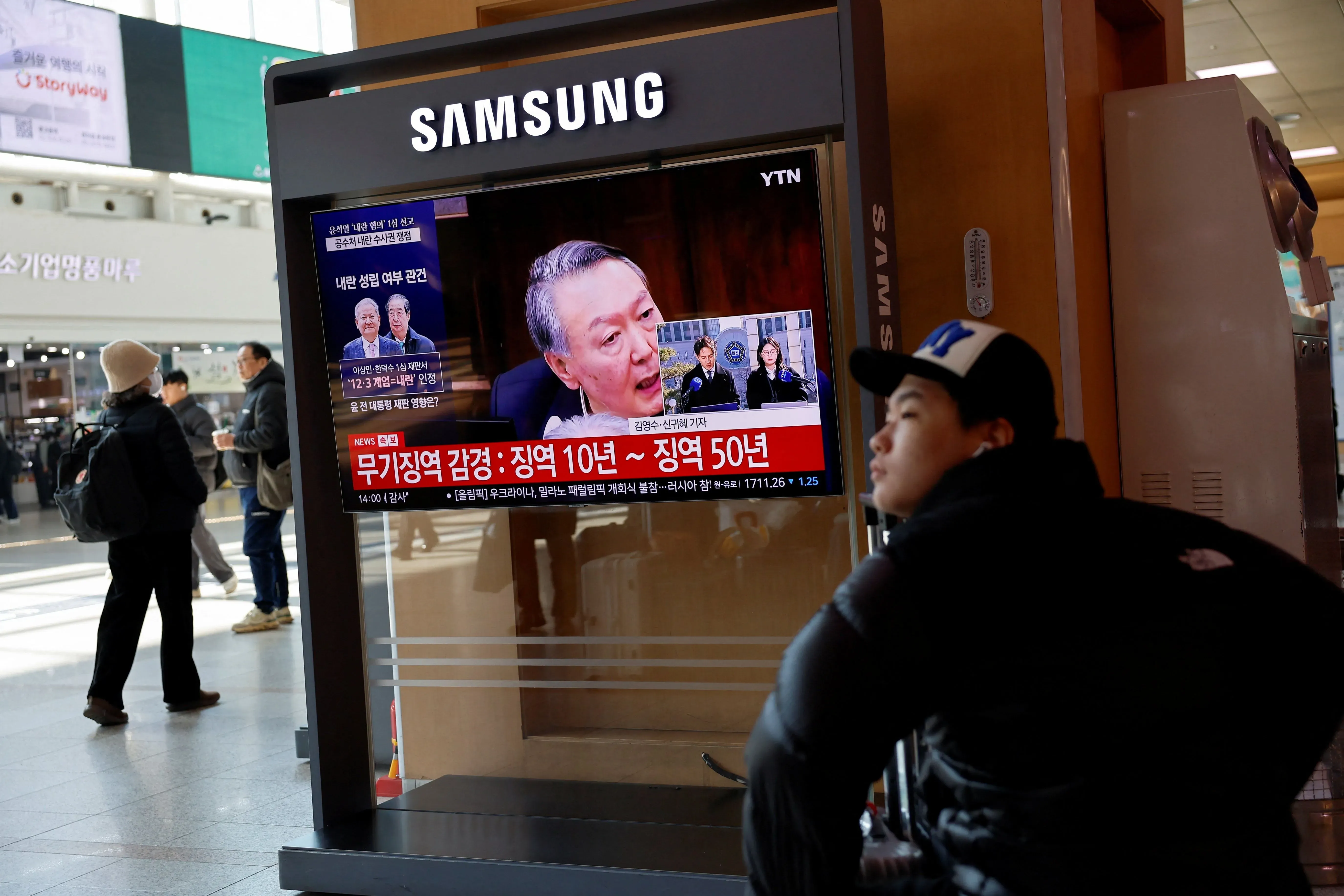 شخصان يشاهدان شاشة عرض في محطة، تعرض أخبارًا عن الرئيس الكوري الجنوبي السابق يون سوك يول وحكمه بالسجن مدى الحياة بتهمة قيادة تمرد.