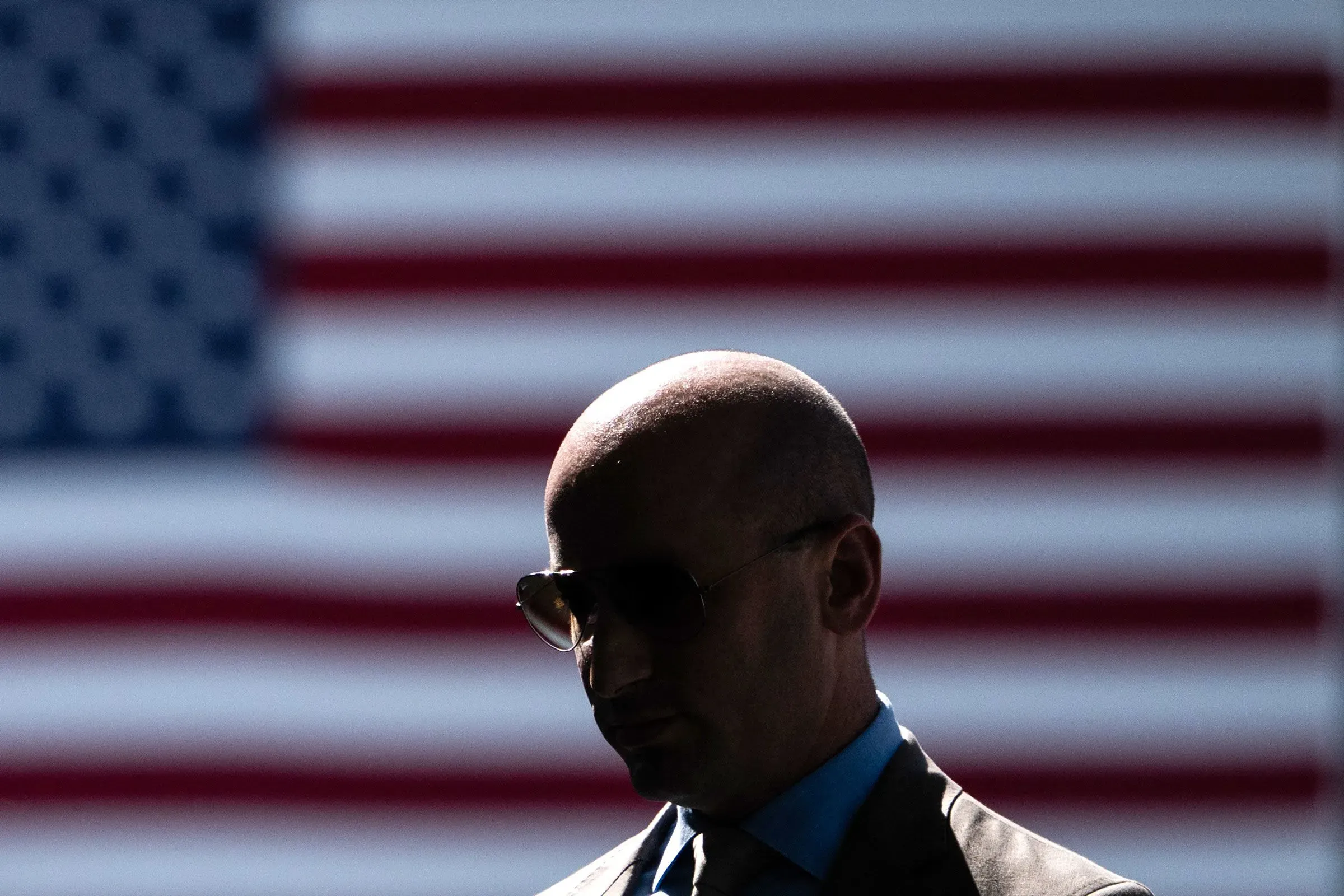 ظل رجل يرتدي نظارات شمسية بدلاً من الخلفية المميزة لعلم الولايات المتحدة، مما يبرز التوترات السياسية في البلاد.