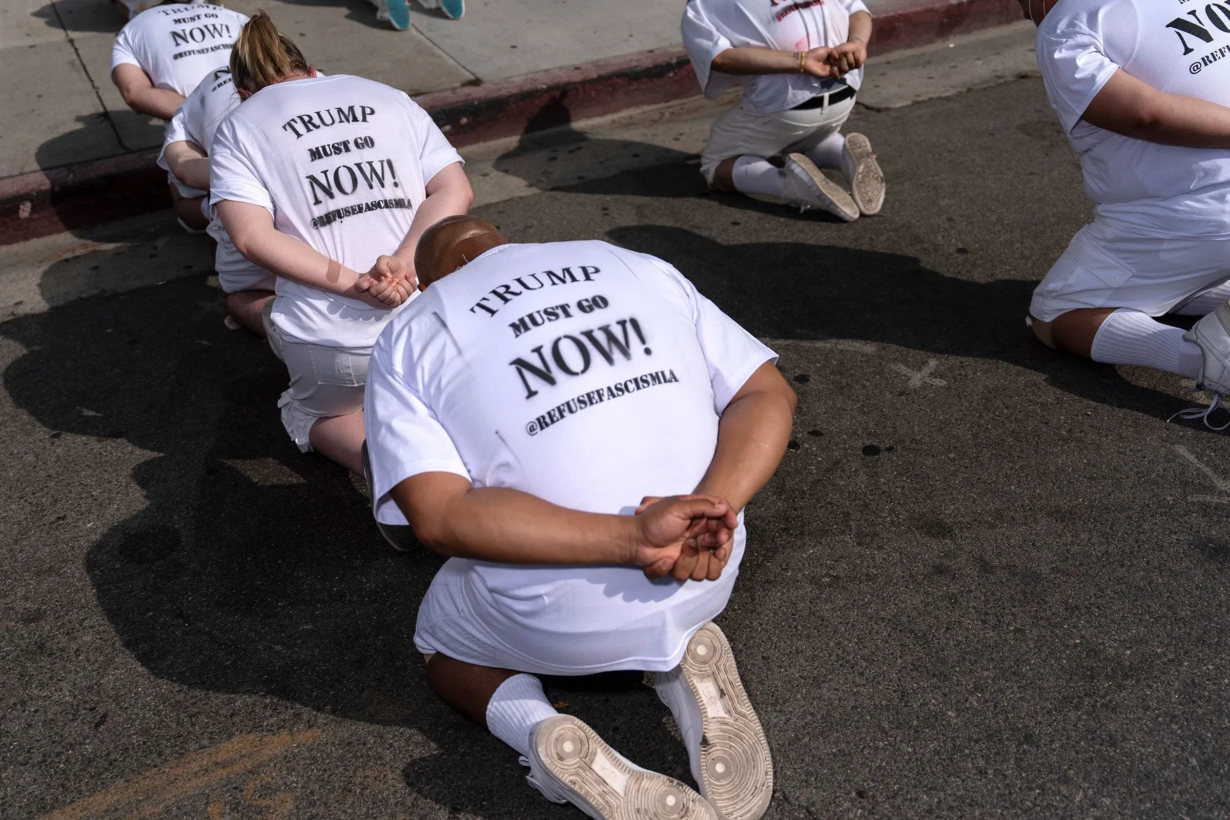 احتجاجات تتضمن مجموعة من الأشخاص يرتدون قمصانًا بيضاء مكتوب عليها "ترامب يجب أن يرحل الآن!"، مع تظاهرهم بالركوع في الشارع.