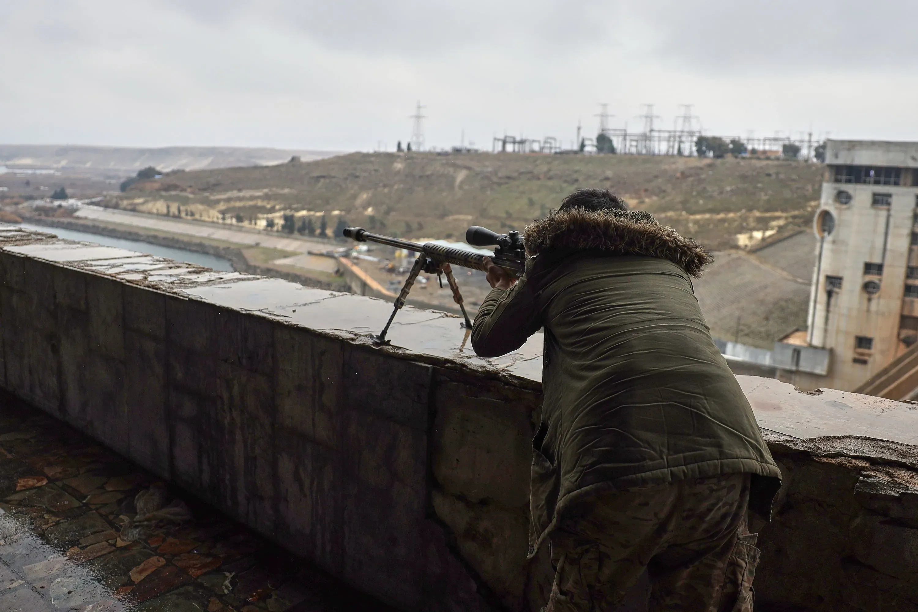 مقاتل من الجيش السوري يتخذ وضعية الاستعداد على حافة مبنى، يراقب المنطقة المحيطة بسد على نهر الفرات، وسط أجواء عسكرية.