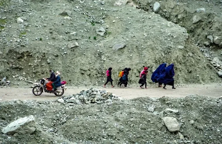 مجموعة من الأشخاص يسيرون على طريق جبلي ضيق في منطقة نائية، بينما يمر رجل على دراجة نارية بجانبهم.