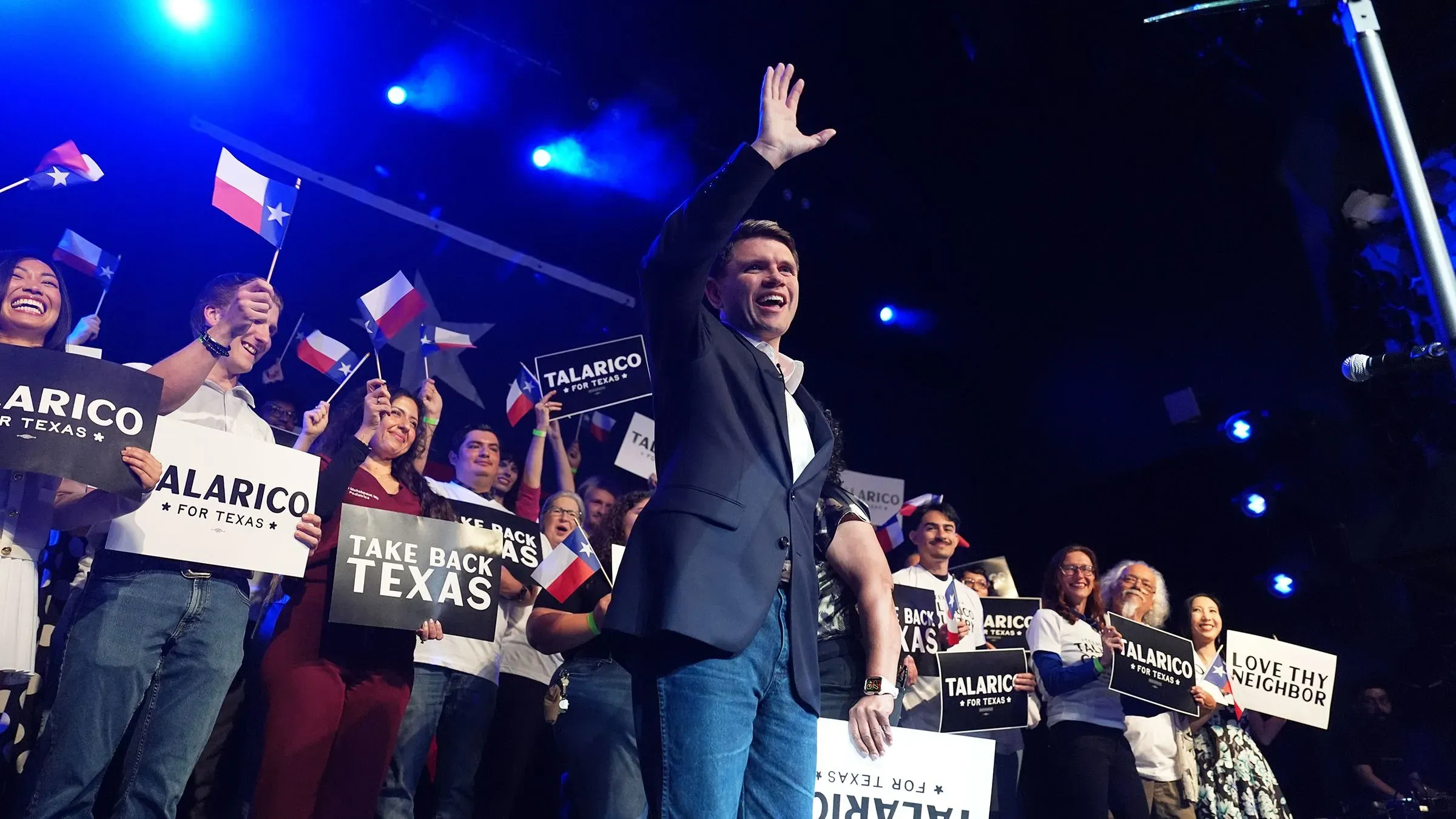 جيمس تالاريكو يحيي الحضور في حدث انتخابي بولاية تكساس، محاطًا بمؤيدين يحملون لافتات وأعلام الولاية، مع أجواء حماسية.