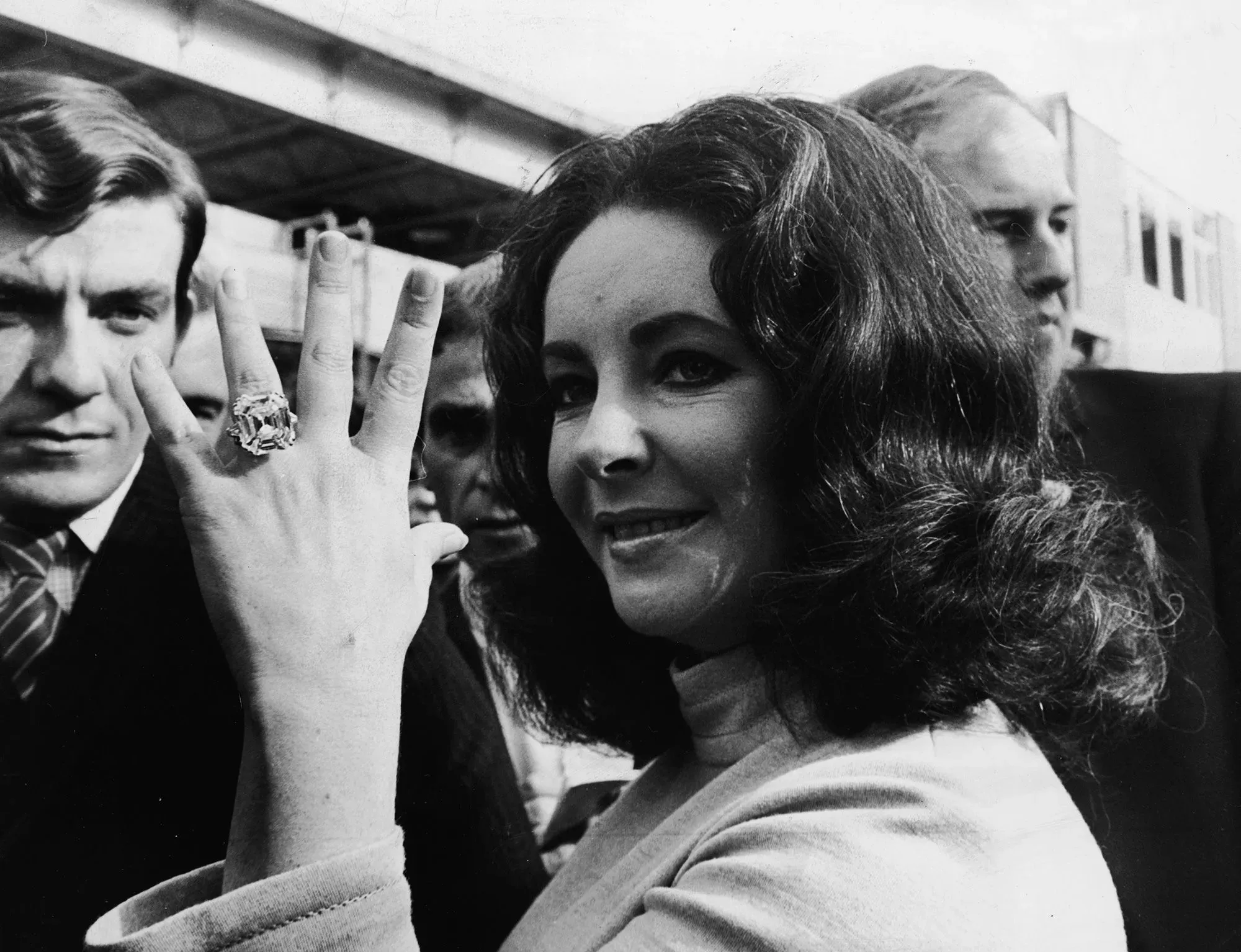 إليزابيث تايلور تظهر بفخر خاتم خطوبتها الكبير مع ماسة بارزة، محاطة بجمهور. تعكس الصورة توجهات حديثة في خواتم الخطوبة.