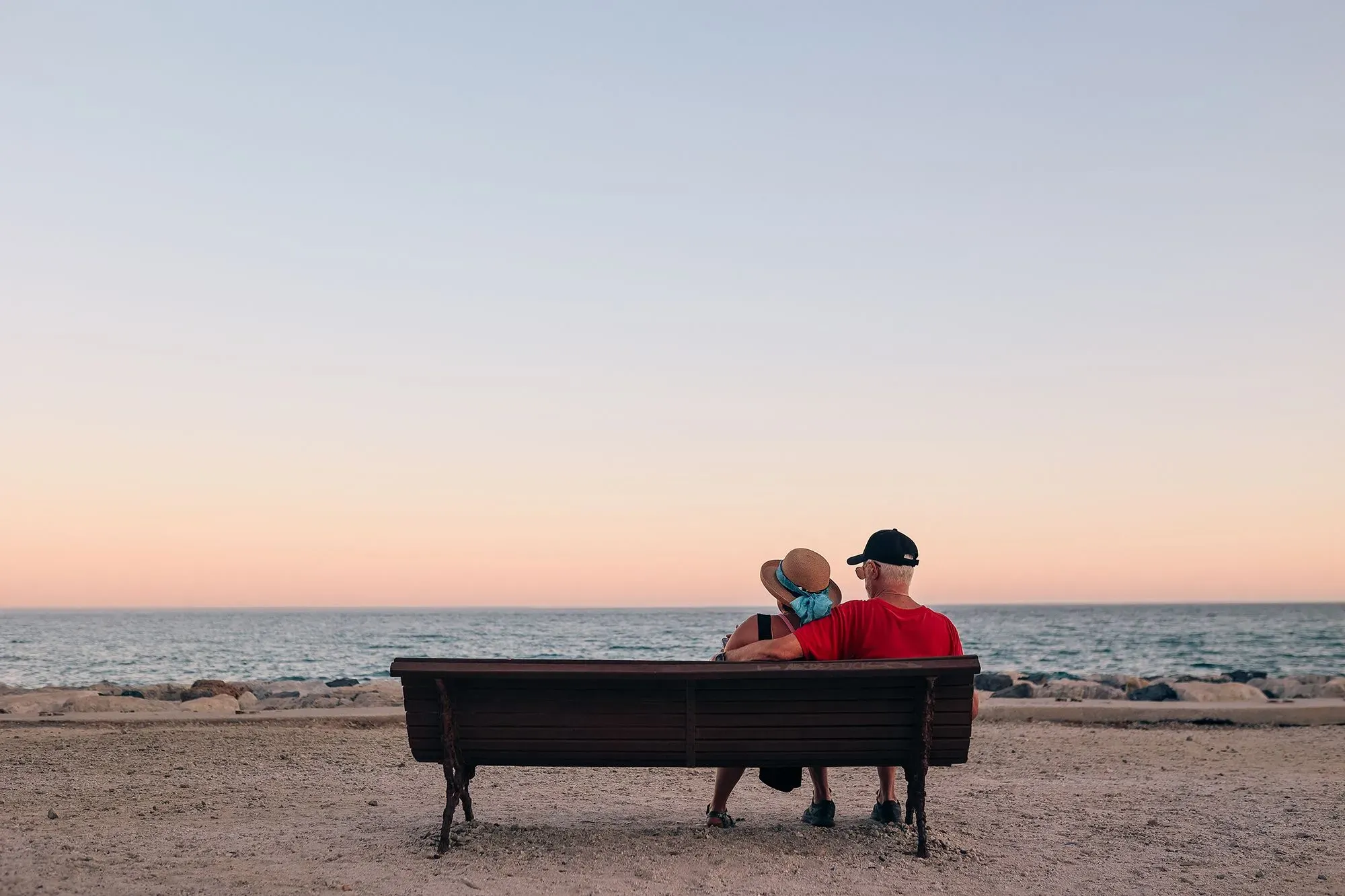 زوجان مسنّان يجلسان على مقعد خشبي أمام البحر، يستمتعان بغروب الشمس، مما يعكس فكرة التقاعد في الخارج وجودة الحياة.