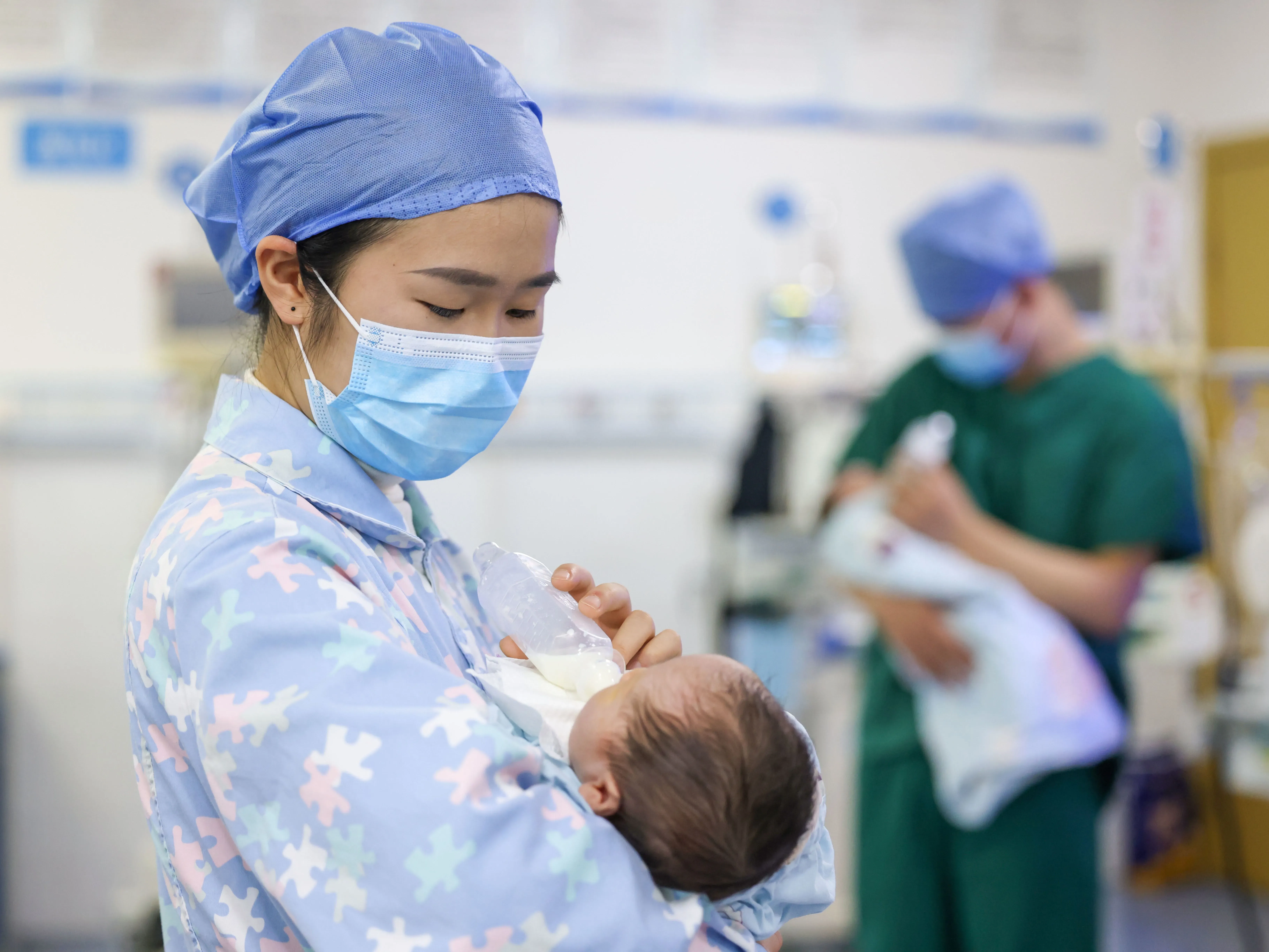مربية ترتدي زيًا طبيًا وتحتضن طفلًا حديث الولادة في مستشفى، بينما يظهر طبيب في الخلفية. تعكس الصورة التحديات في رعاية الأطفال في الصين.