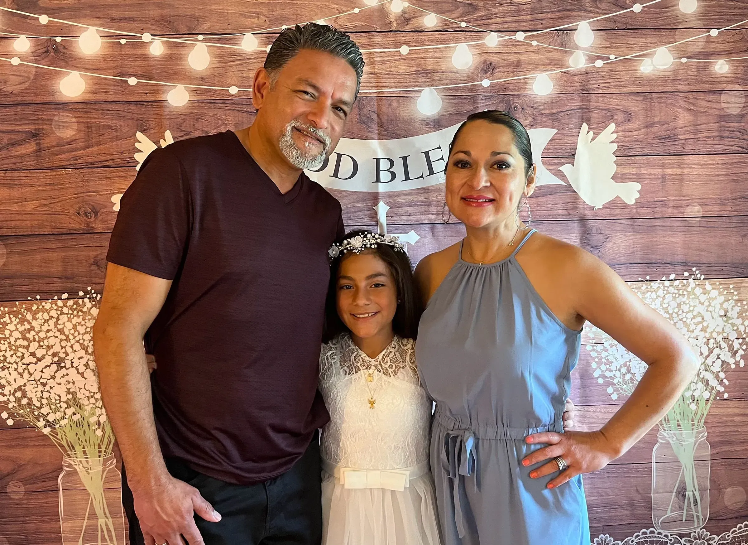 عائلة مكونة من ثلاثة أفراد، تضم شخصين بالغين وطفلة ترتدي فستانًا أبيض، في خلفية مزينة بأضواء وعبارات احتفالية.