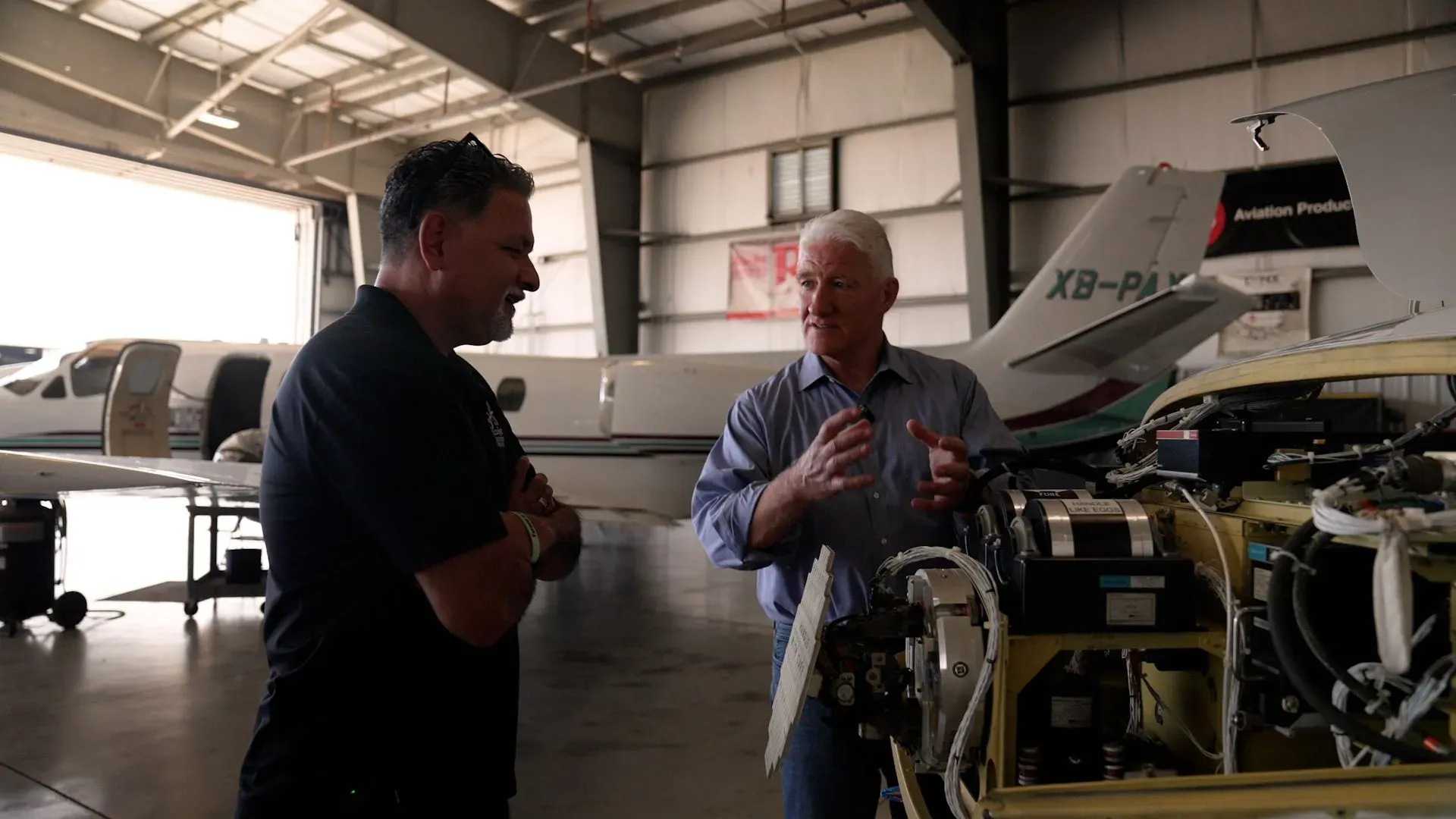 رجلان يتحدثان داخل حظيرة طائرات، أحدهما يشرح تفاصيل فحص وإصلاح طائرة، بينما خلفهما طائرتان في وضعية انتظار.