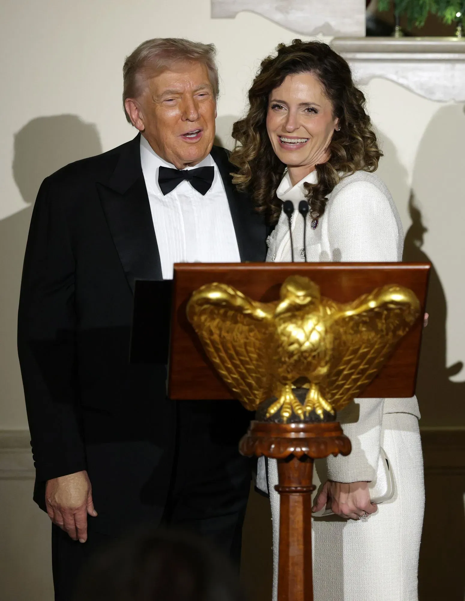 صورة لدونالد ترامب مبتسمًا بجانب امرأة ترتدي فستانًا أبيض، أثناء حدث رسمي، مع وجود شعار نسر خلفهما.