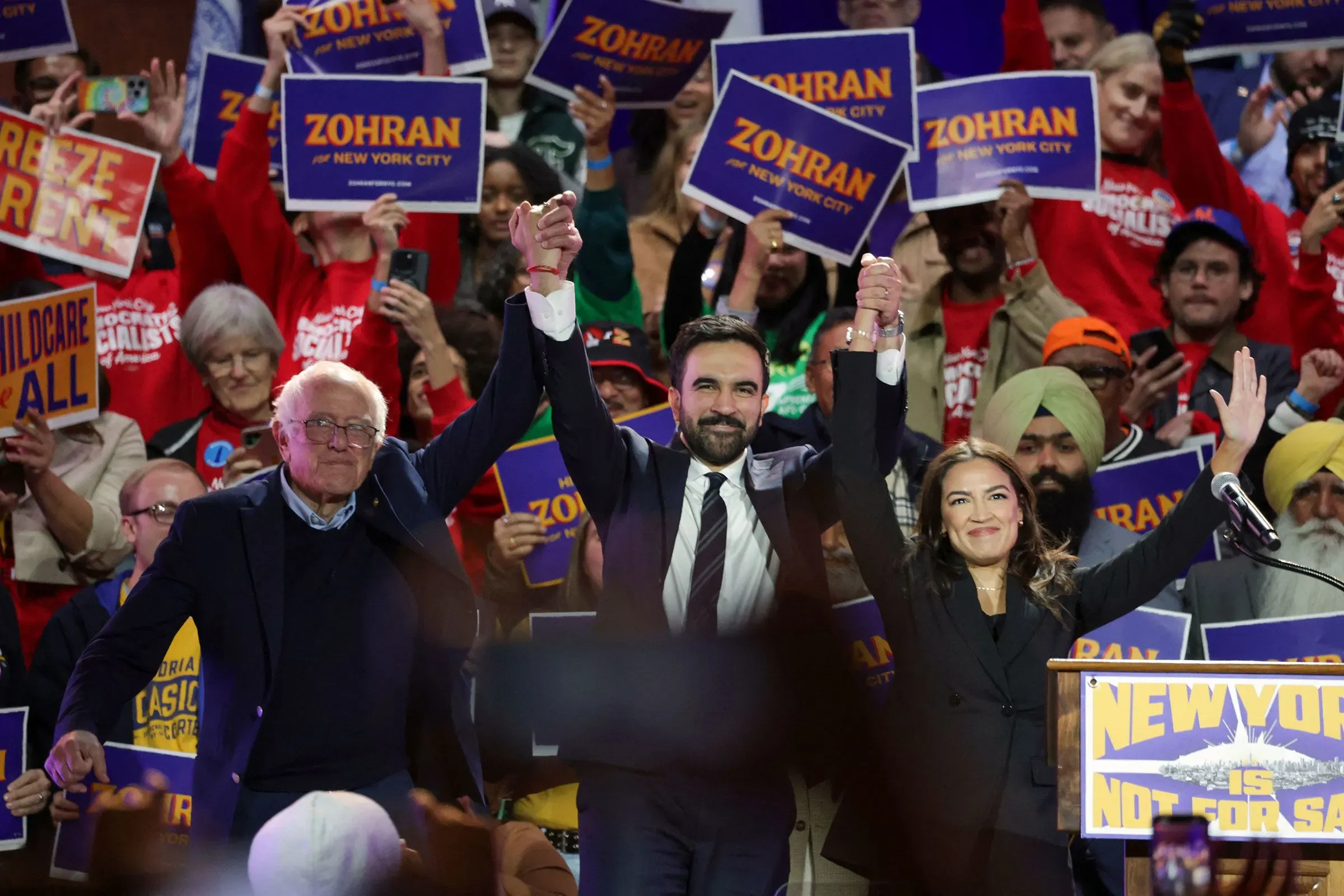 تجمع حاشد لدعم المرشح زهران ممداني، حيث يظهر برفقة شخصيات سياسية بارزة مثل ألكساندريا أوكاسيو كورتيز وساندرز، وسط لافتات انتخابية.