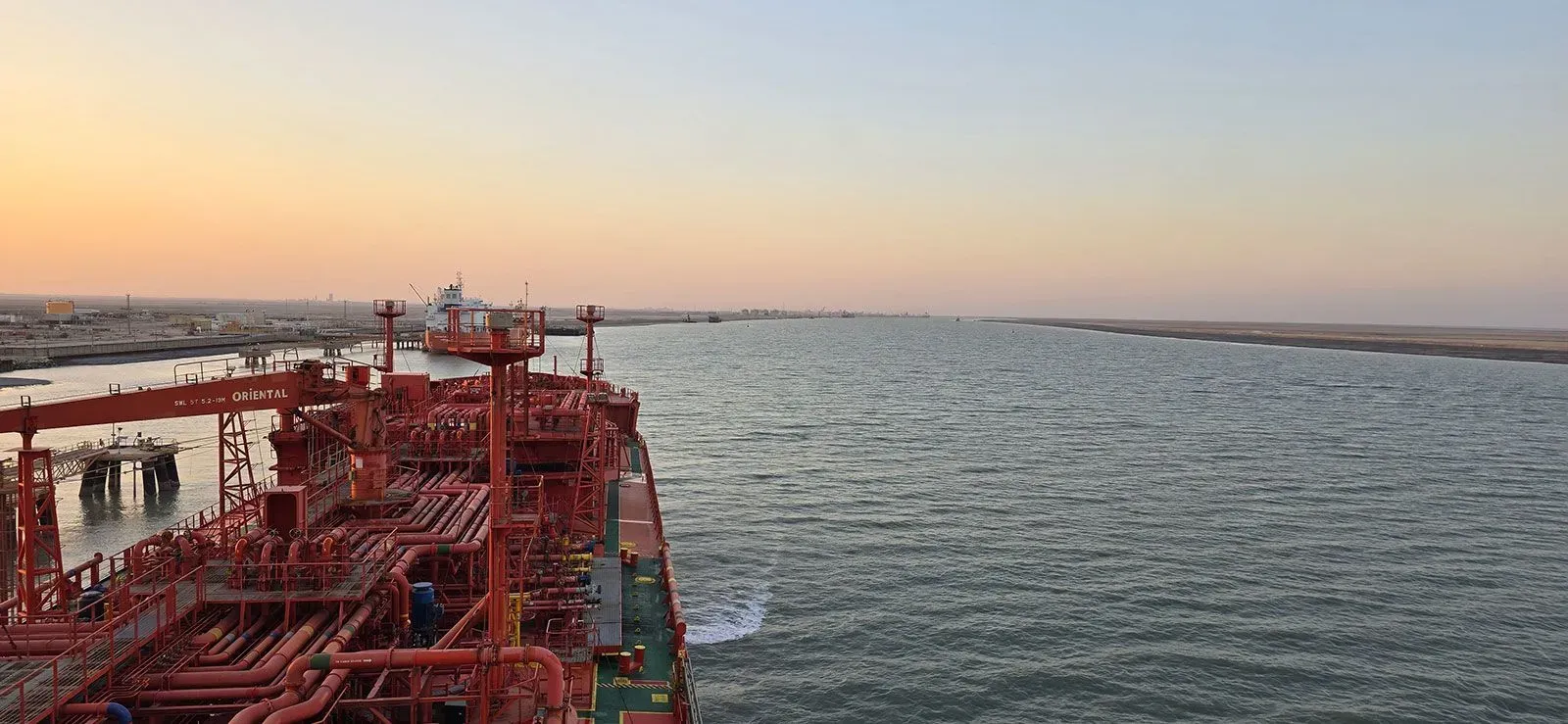 صورة لناقلة نفط حمراء ترسو في مياه الخليج العربي عند غروب الشمس، مع وجود ساحل بعيد في الخلفية، تعكس التوترات الحالية في الشحن البحري.