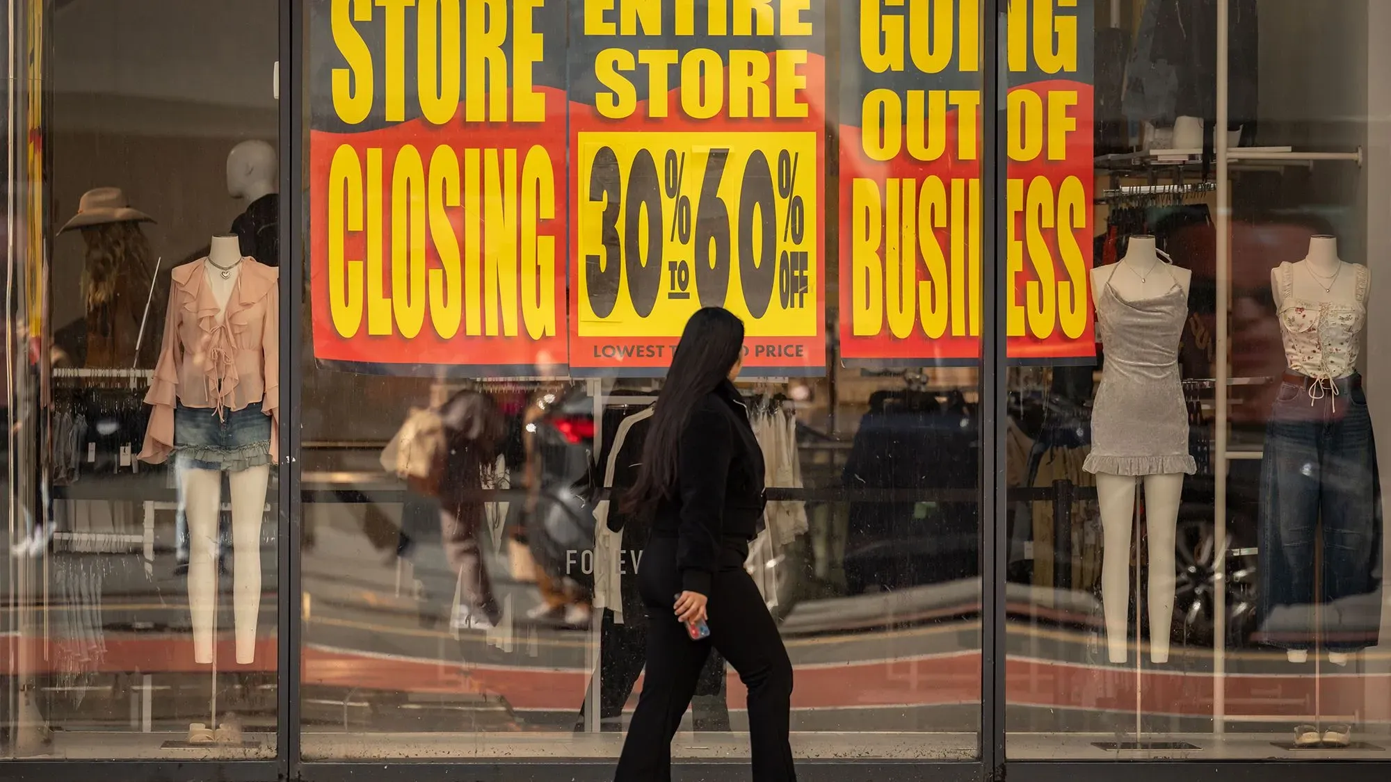 امرأة ترتدي ملابس سوداء تسير أمام واجهة متجر مغلق، مع لافتات كبيرة تشير إلى إغلاق المتجر وتخفيضات تصل إلى 60%.