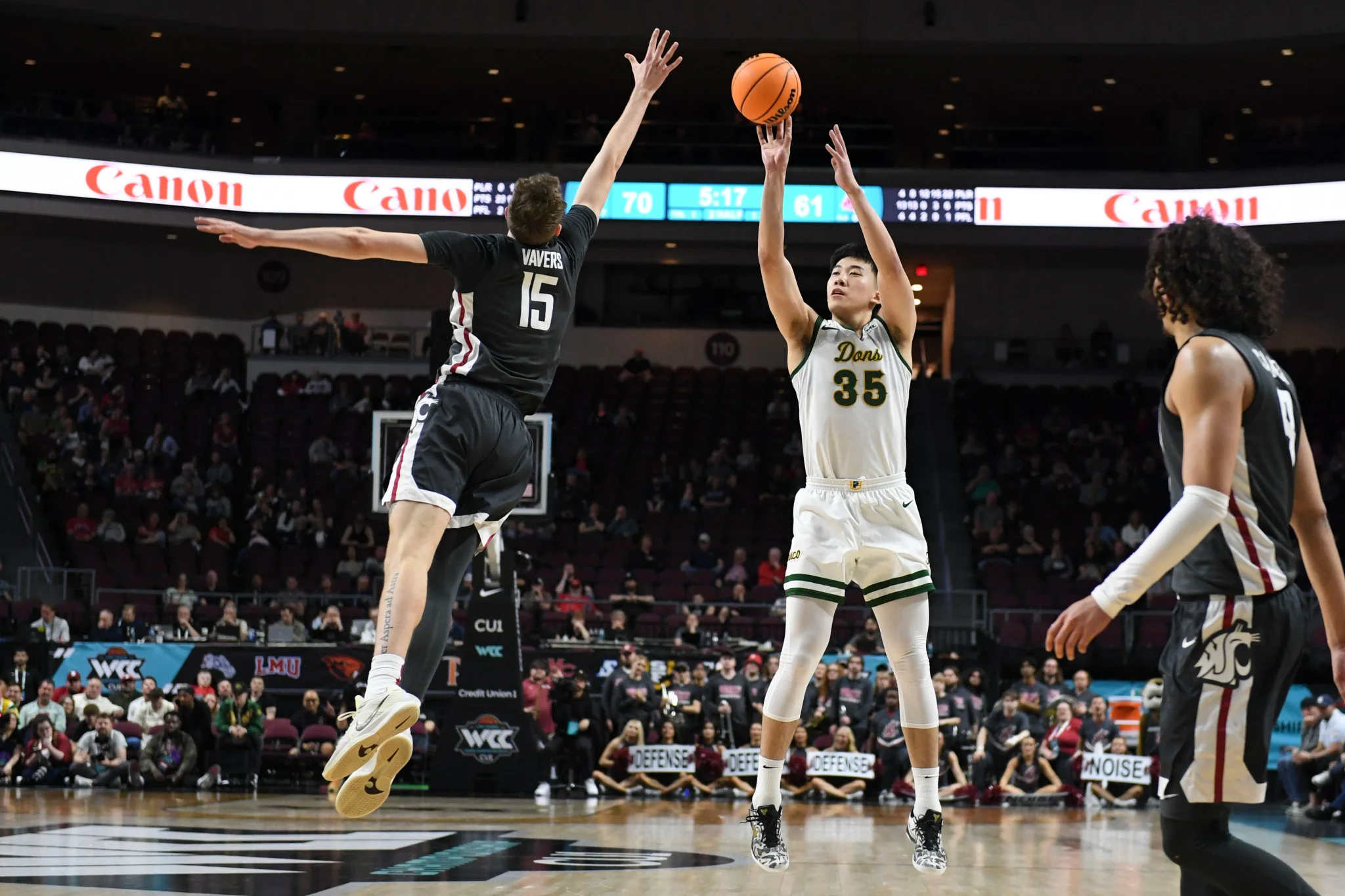 لاعب كرة سلة صيني يرتدي زي فريقه الأبيض رقم 35 يسدد كرة في مباراة NCAA وسط دفاع منافس يرتدي زي أسود، مع جمهور في الخلفية.