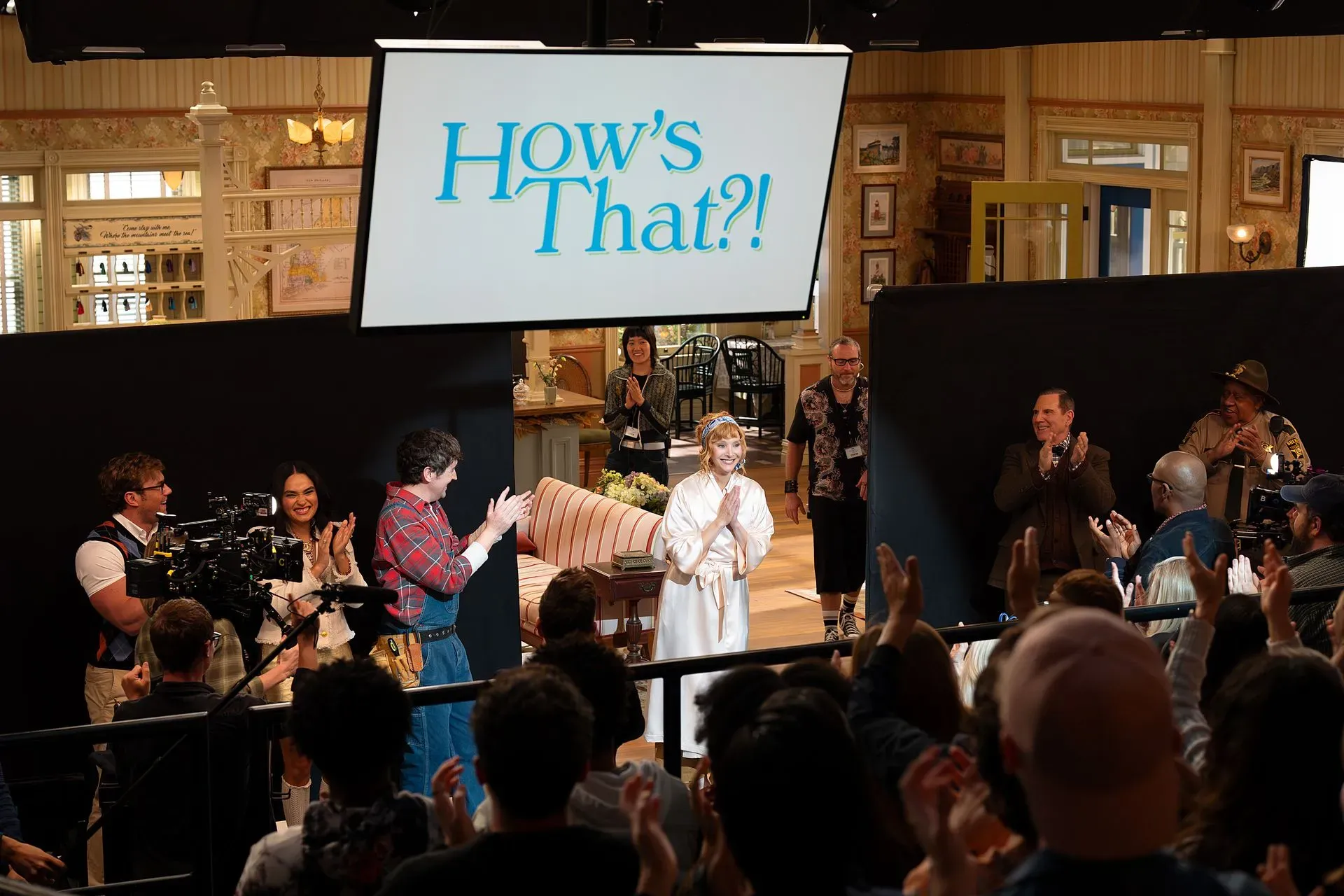 فاليري شيريش، الشخصية الرئيسية في مسلسل "The Comeback"، تتلقى تصفيقاً حاراً أثناء تصوير مشهد جديد، مع لافتة "How's That?!" في الخلفية.