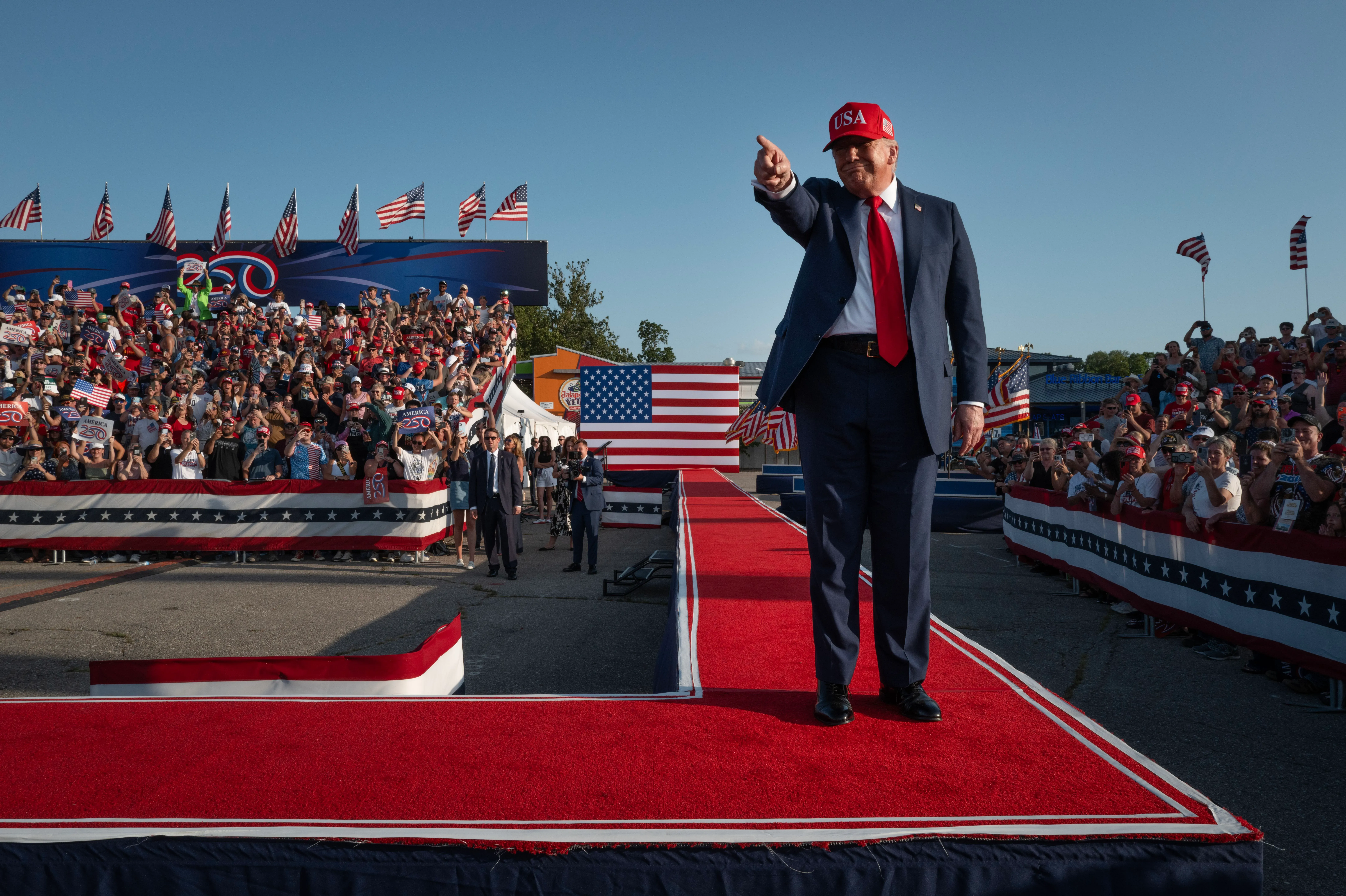ترامب يشير بيده إلى الحشد في تجمع انتخابي، مع وجود أعلام أمريكية خلفه، مما يعكس دعمه المتواصل من قاعدة جمهورية متزايدة الانقسام.