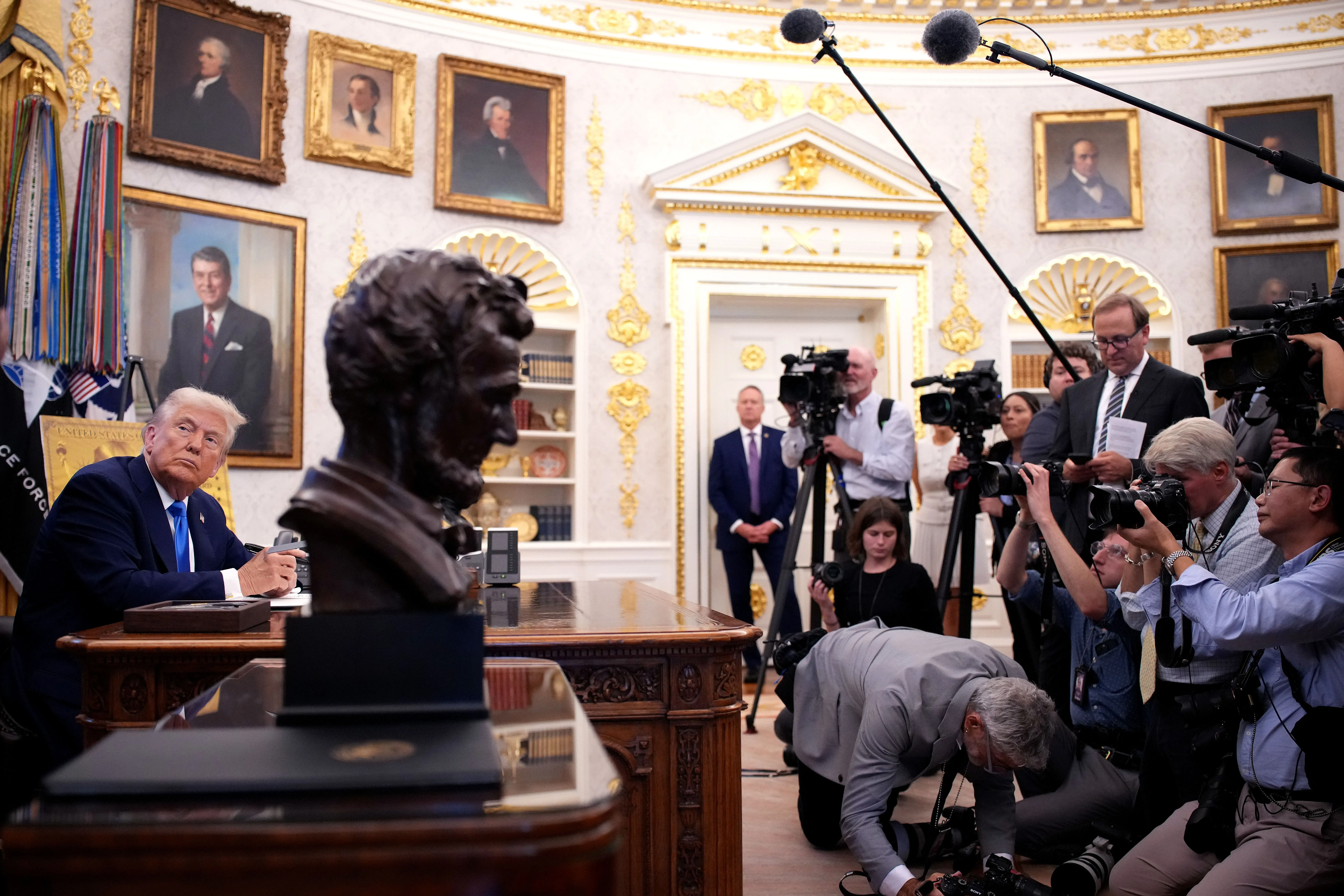 ترامب يجلس في المكتب البيضاوي، مع تمثال نصفي لأبراهام لينكولن أمامه، بينما يحيط به الصحفيون والكاميرات.