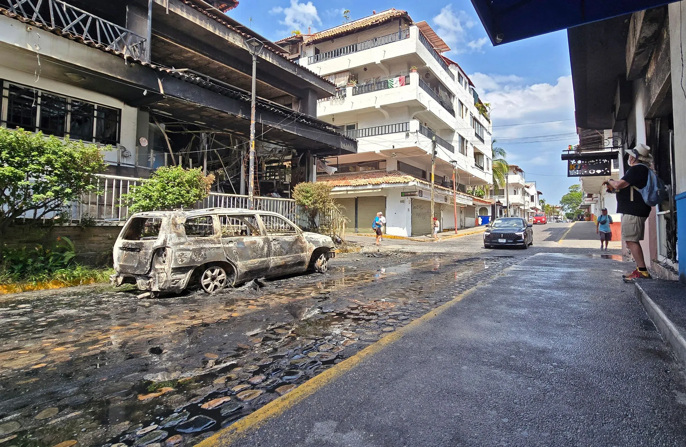 شارع في بويرتو فالارتا يظهر سيارة محترقة وأضرار في المباني، مما يعكس آثار أعمال العنف التي اجتاحت المدينة بعد مقتل زعيم كارتل خاليسكو.