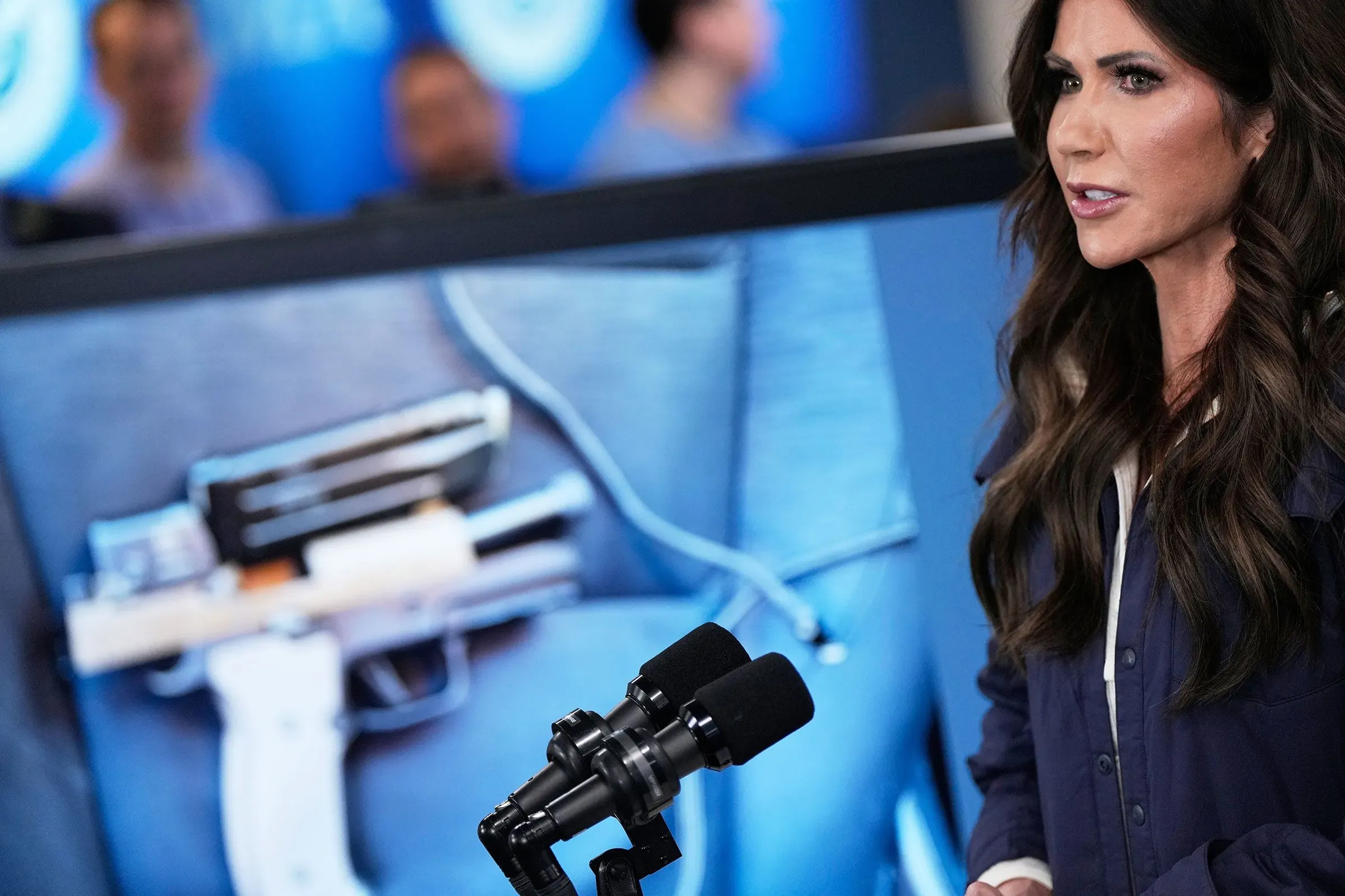 تظهر الصورة كريستي نويم، وزيرة الأمن الداخلي، تتحدث أمام ميكروفونات مع عرض لصورة سلاح ناري خلفها، في سياق مناقشة قضايا الهجرة.