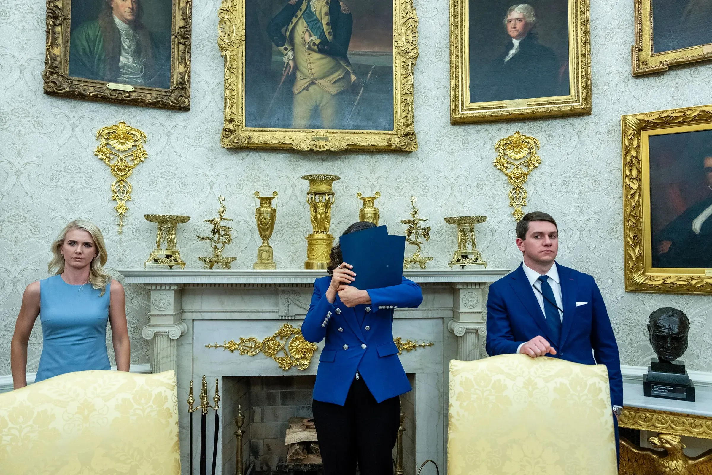 مجموعة من الأشخاص يقفون في المكتب البيضاوي، مع وجود لوحات تاريخية على الجدران. يظهر شخص يحمل ورقة ويغطي وجهه، بينما يرتدي الآخرون ملابس رسمية.