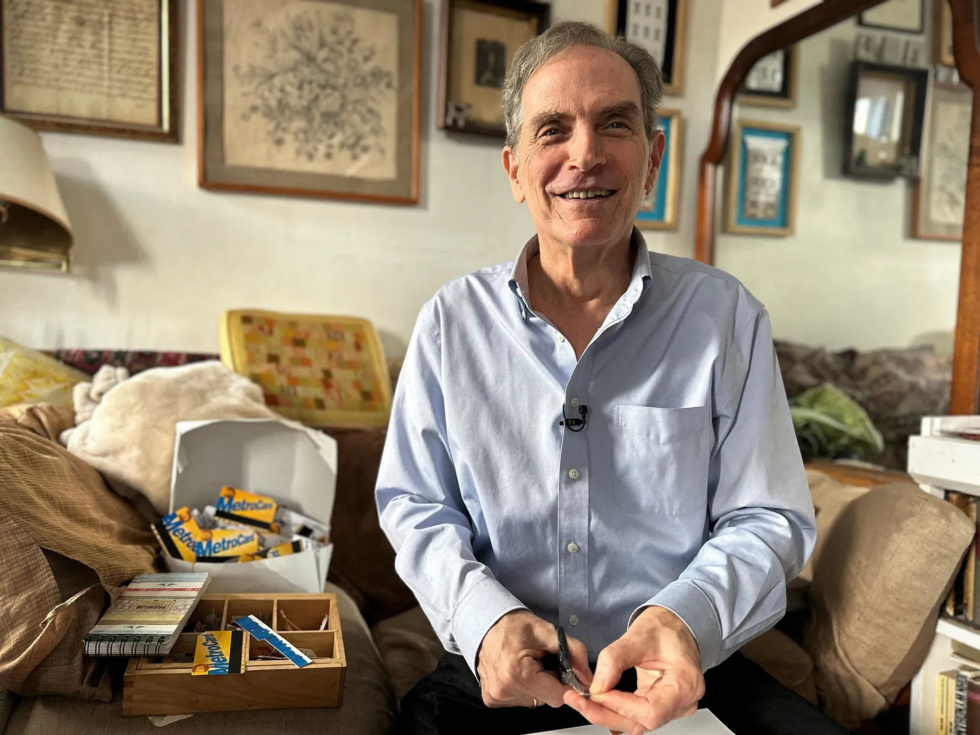 رجل مبتسم يجلس في غرفة معيشته، محاط ببطاقات مترو قديمة ومجموعة من التذكارات، مما يعكس شغفه بجمع بطاقات المترو في نيويورك.