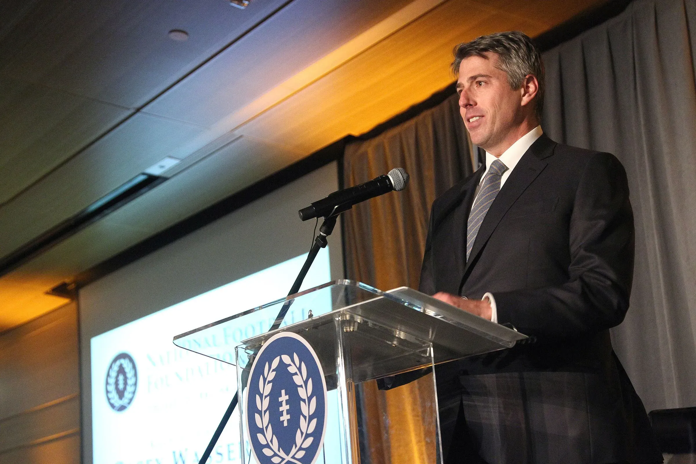 كيسي واسرمان يتحدث في حدث رسمي، مع خلفية تتضمن شعار المؤسسة. يواجه ضغوطًا بسبب ارتباطاته مع غيسلين ماكسويل.