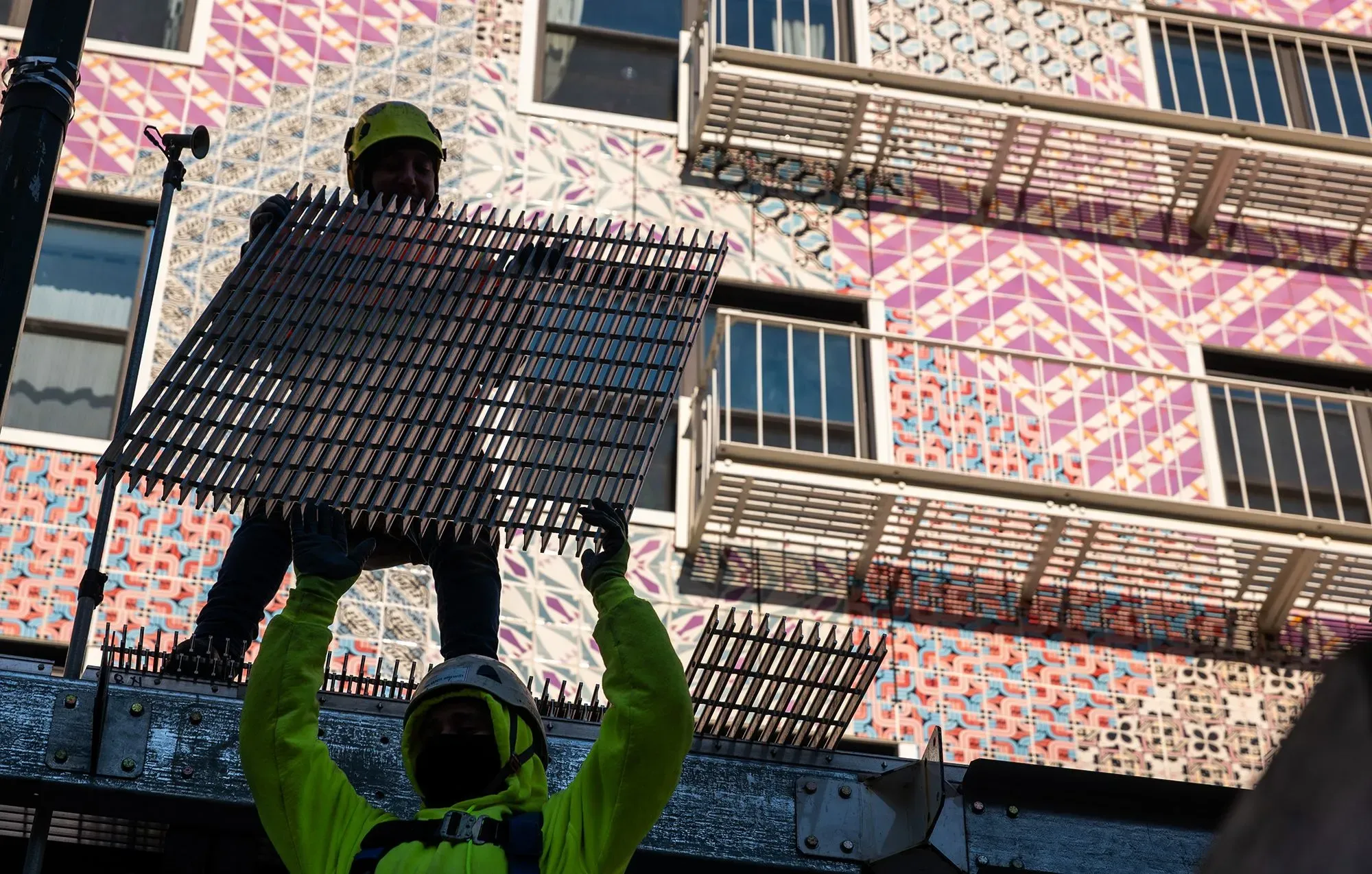 عمال بناء يحملون لوحًا في موقع إنشاءات، مع خلفية ملونة لجدران المبنى، مما يعكس النشاط الاقتصادي في السوق.