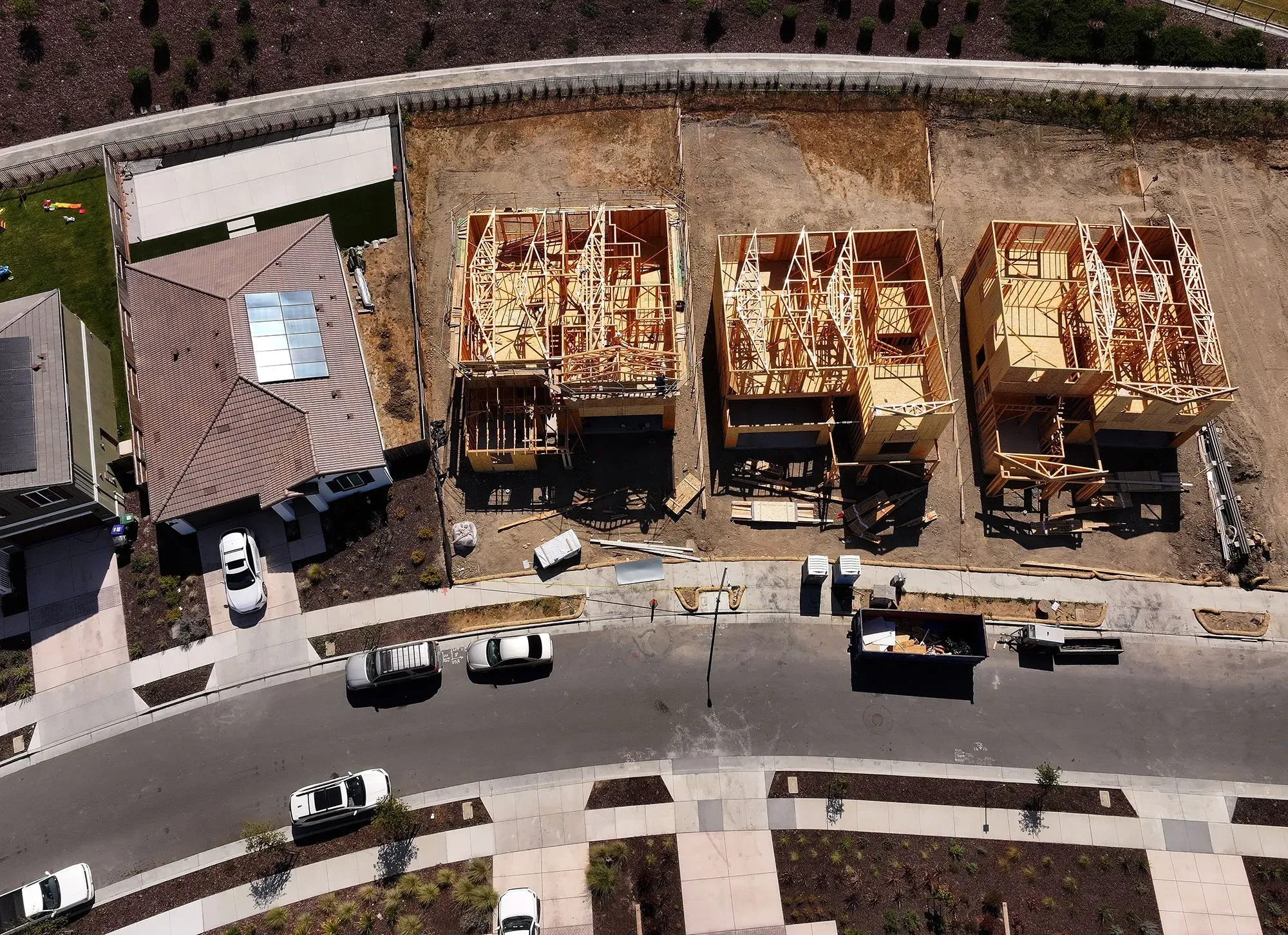 موقع بناء منازل جديدة يظهر ثلاث هياكل خشبية في مرحلة البناء، مع سيارات متوقفة في الشارع المجاور.