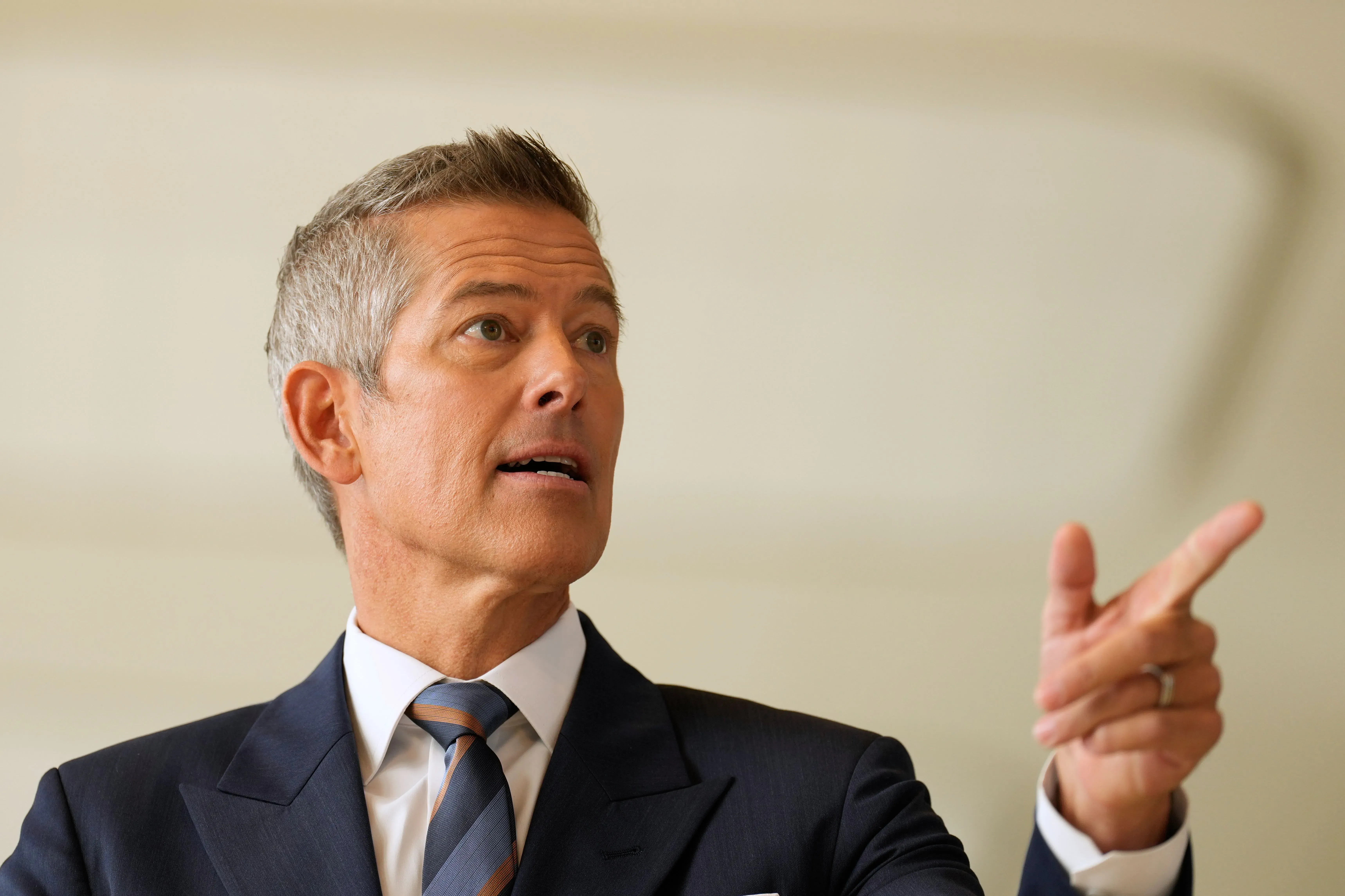 رجل يرتدي بدلة رسمية يتحدث بجدية، مشيرًا بيده، في مؤتمر صحفي حول تأثير الإغلاق الحكومي على مراقبي الحركة الجوية.