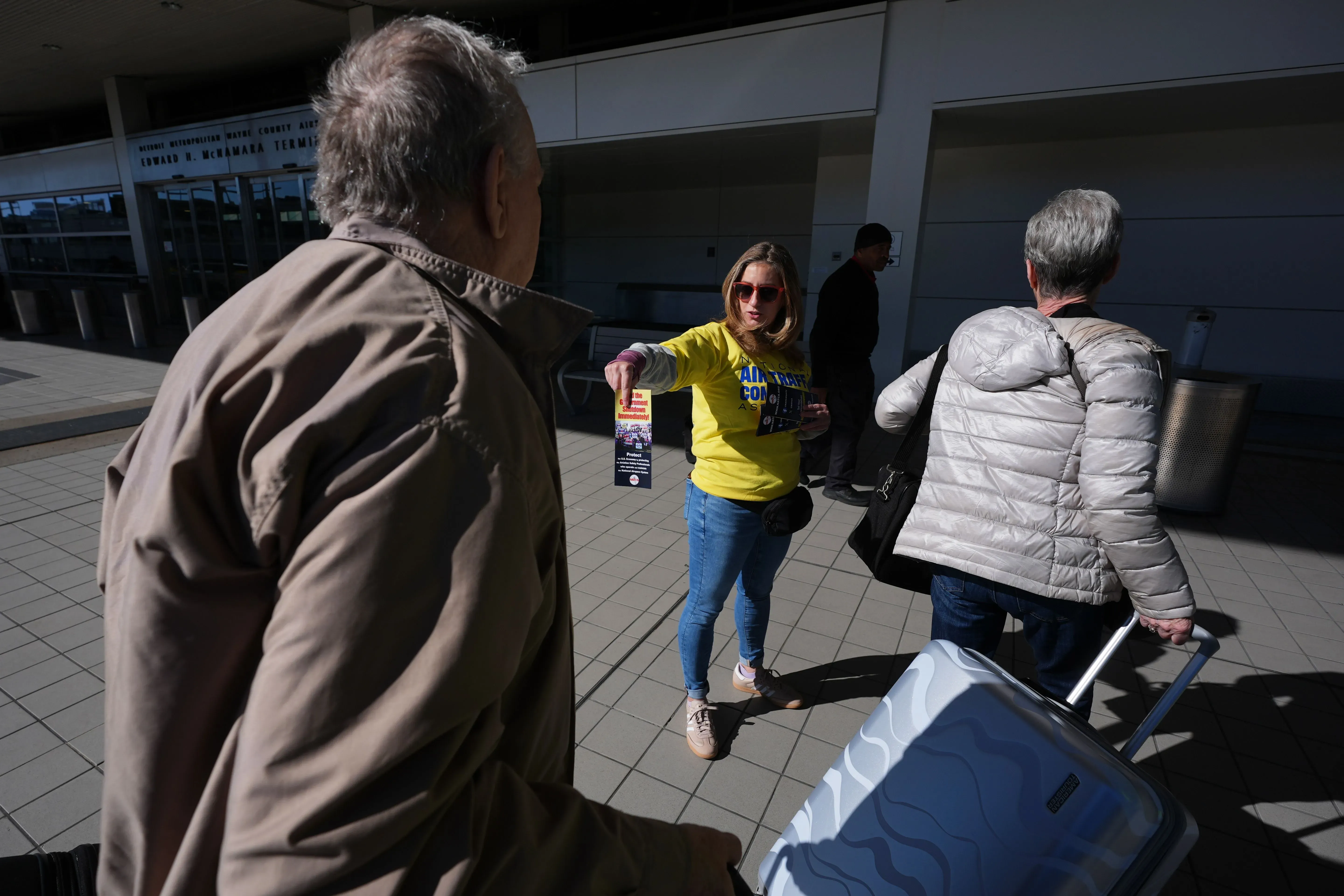 امرأة ترتدي قميصًا أصفر، توزع منشورات في مطار، بينما يتجه المسافرون نحو مكاتب تسجيل الوصول، مما يعكس تأثير الإغلاق الحكومي على خدمات الطيران.