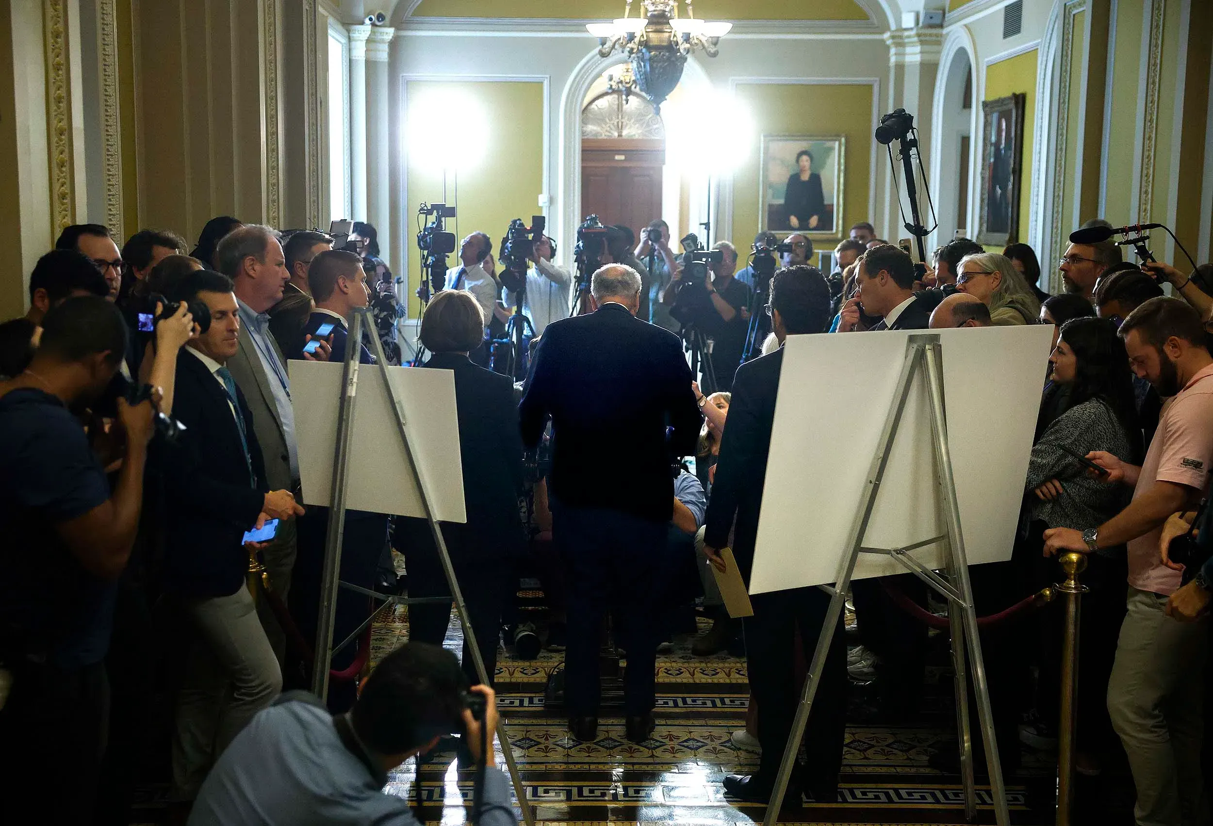 ازدحام صحفي في ممر الكونغرس، مع وجود شخصيات سياسية بارزة في المقدمة، مما يعكس التوترات السياسية المتعلقة بالإغلاق الحكومي.