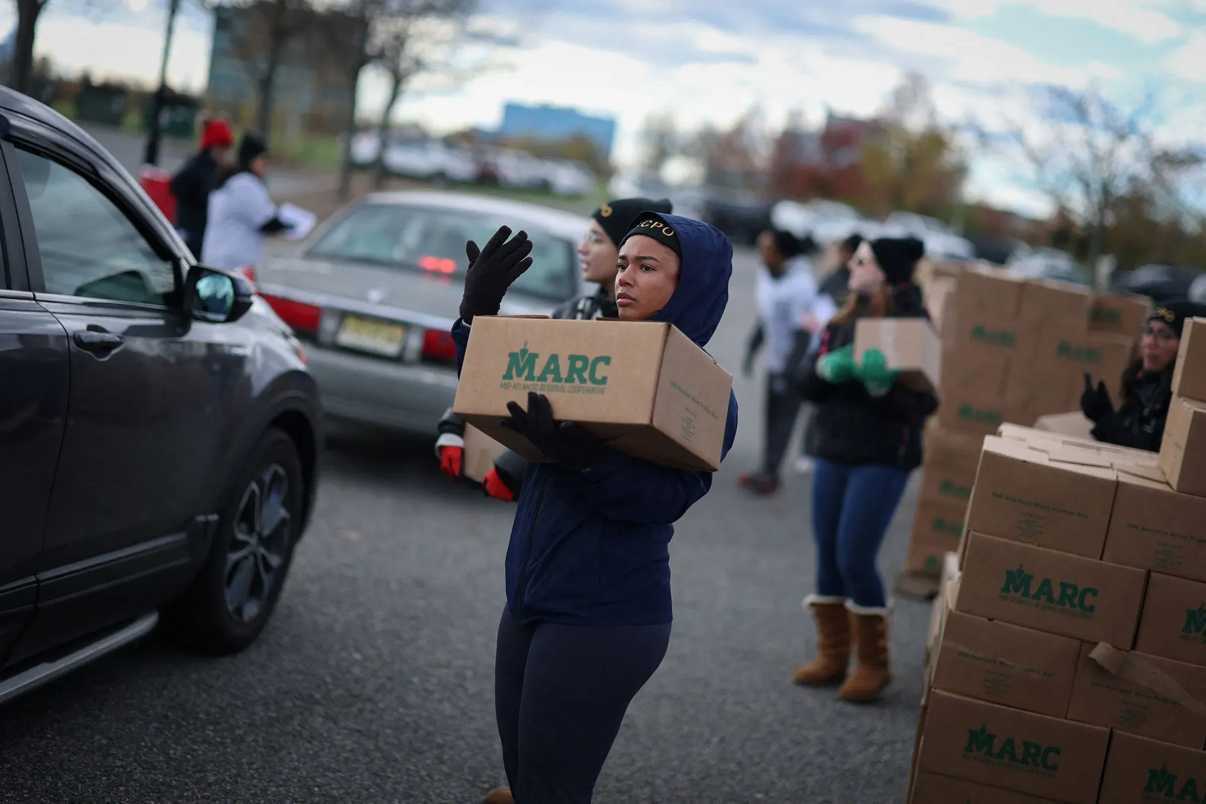 موظفة تحمل صندوقًا في حدث توزيع للمواد الغذائية، مع وجود سيارات وأشخاص آخرين في الخلفية، مما يعكس جهود الدعم المجتمعي خلال الإغلاق الحكومي.