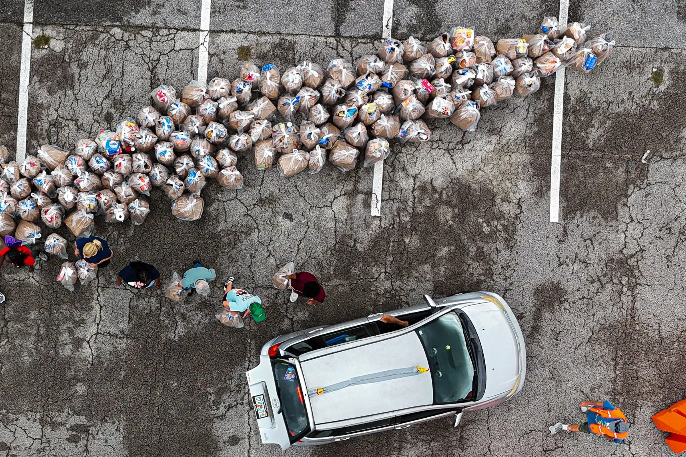طوابير من الأشخاص يتلقون المساعدات الغذائية من أكياس مملوءة بالطعام، مع سيارة متوقفة بالقرب من المساعدات في موقف سيارات.