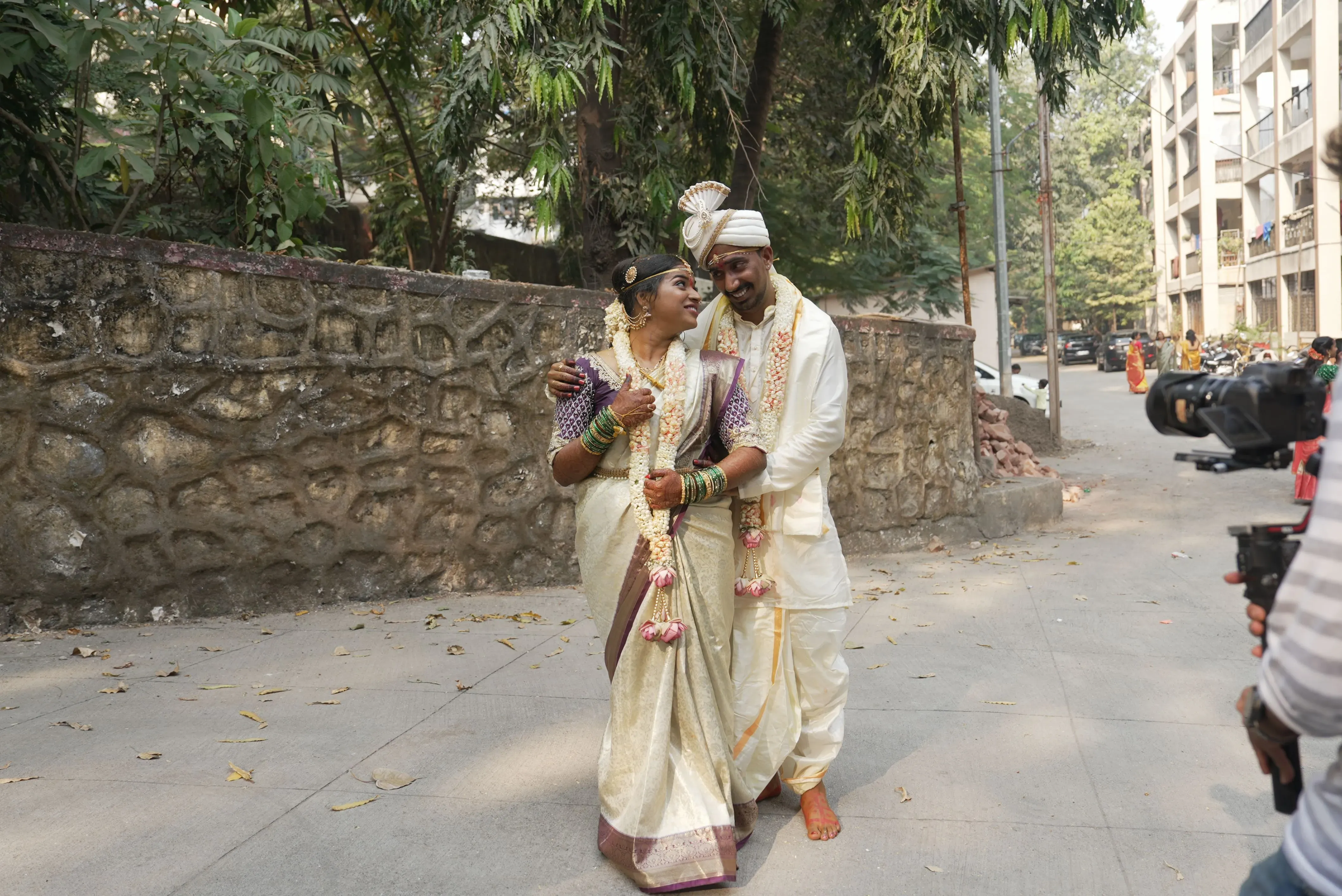 عروس وعريس مبتسمان يرتديان ملابس تقليدية في حفل زفاف، محاطان بأجواء احتفالية في مومباي، مع تفاصيل دقيقة من الزهور والديكور.