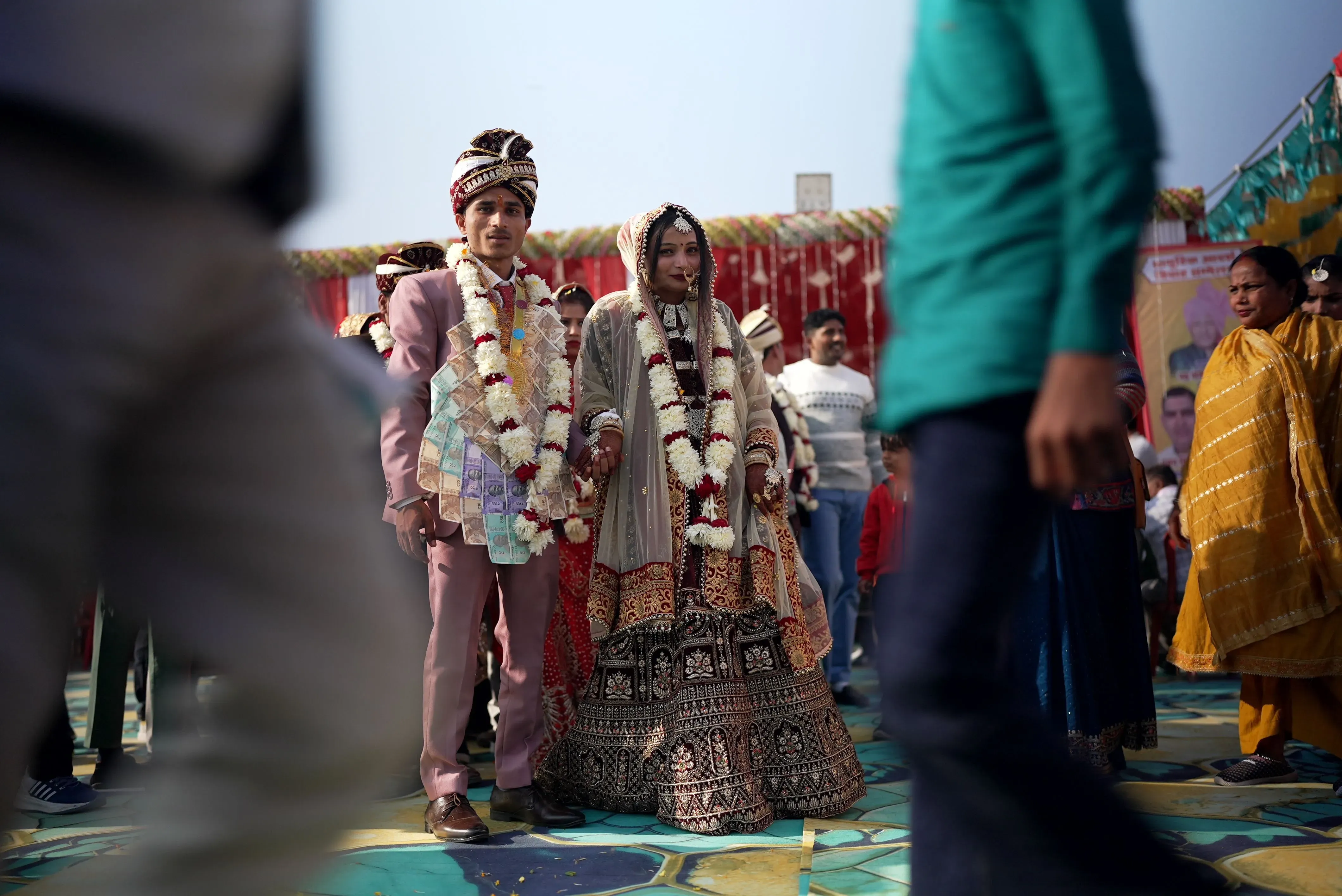 عروس وعريس يرتديان ملابس تقليدية مزينة بالزهور، يتوسطان حفل زفاف مزدحم في الهند، مع ضيوف يتجولون حولهما.