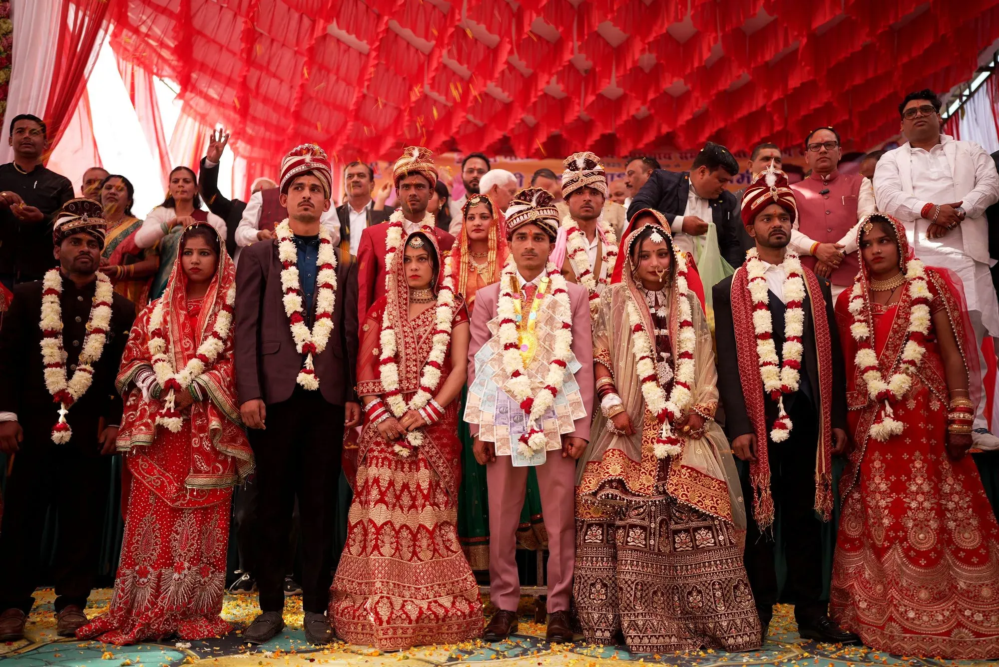 مجموعة من الأزواج في حفل زفاف تقليدي في الهند، يرتدون أزياء ملونة وزهور، مع خلفية مزينة بأقمشة حمراء.