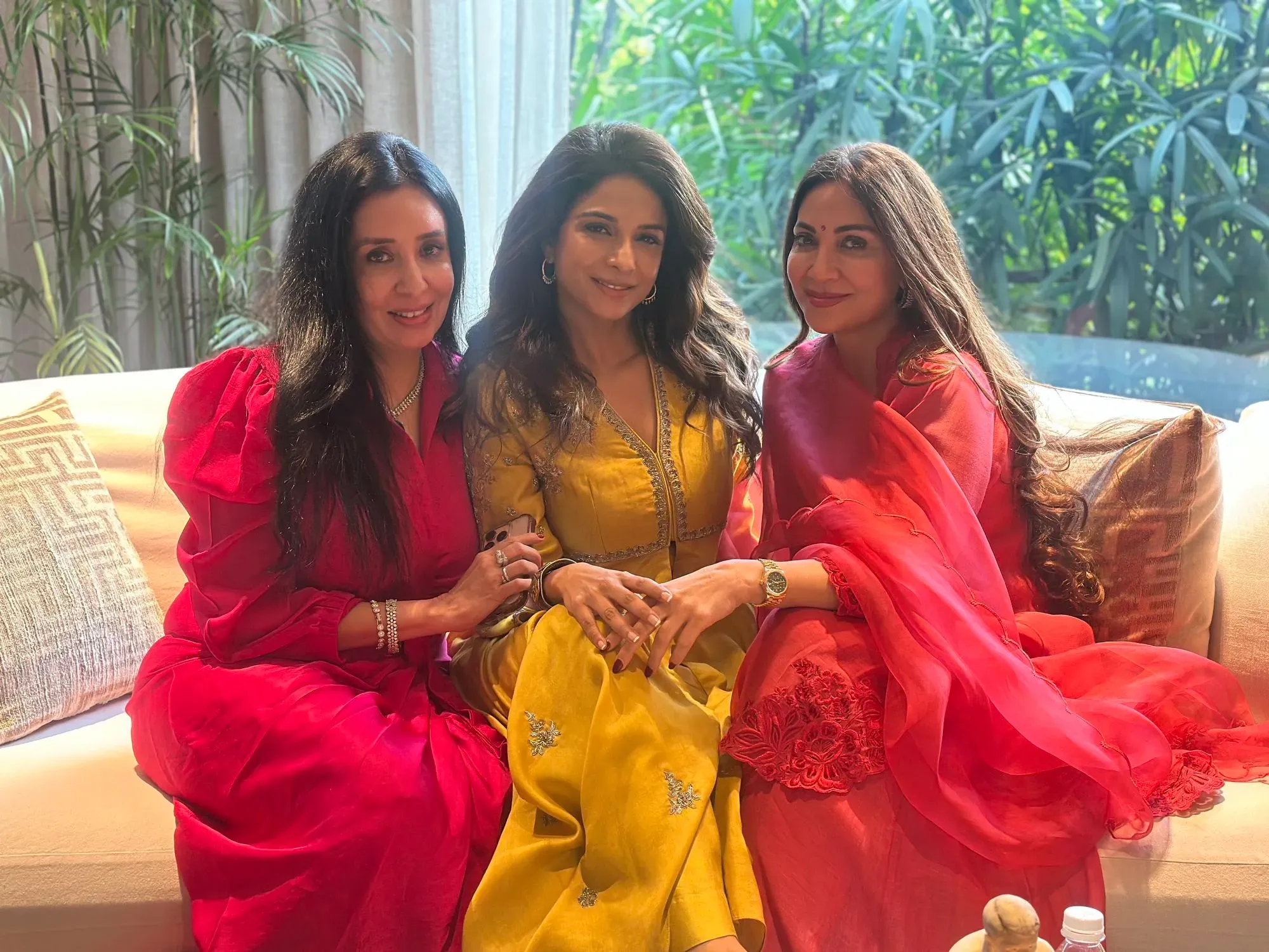 ثلاث نساء يجلسن معًا على أريكة، مرتديات ملابس ملونة، حيث تبتسم إحداهن في وسط الصورة. خلفهن نباتات خضراء تضفي جوًا مريحًا.