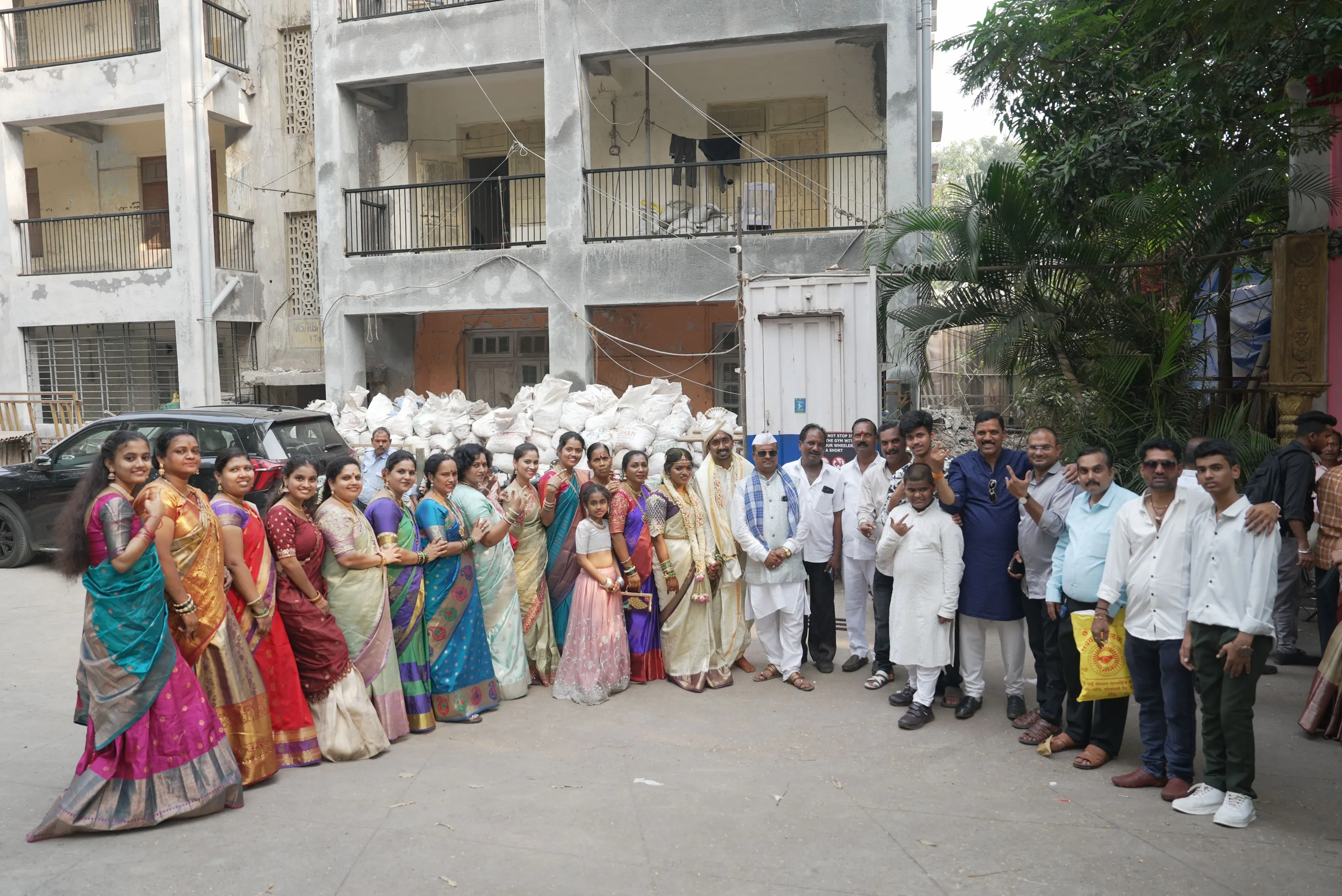 مجموعة من الضيوف يرتدون ملابس تقليدية ملونة، يتجمعون في موقع حفل زفاف في مومباي، مع خلفية لمبانٍ بسيطة وأكياس من المواد.