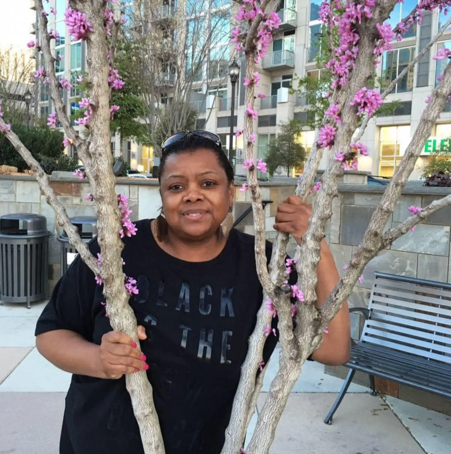 امرأة مبتسمة تحمل أغصان شجرة مزهرة، تظهر في خلفية حضرية مع مبانٍ حديثة. تعكس الصورة لحظة من الفرح والذكريات العائلية.