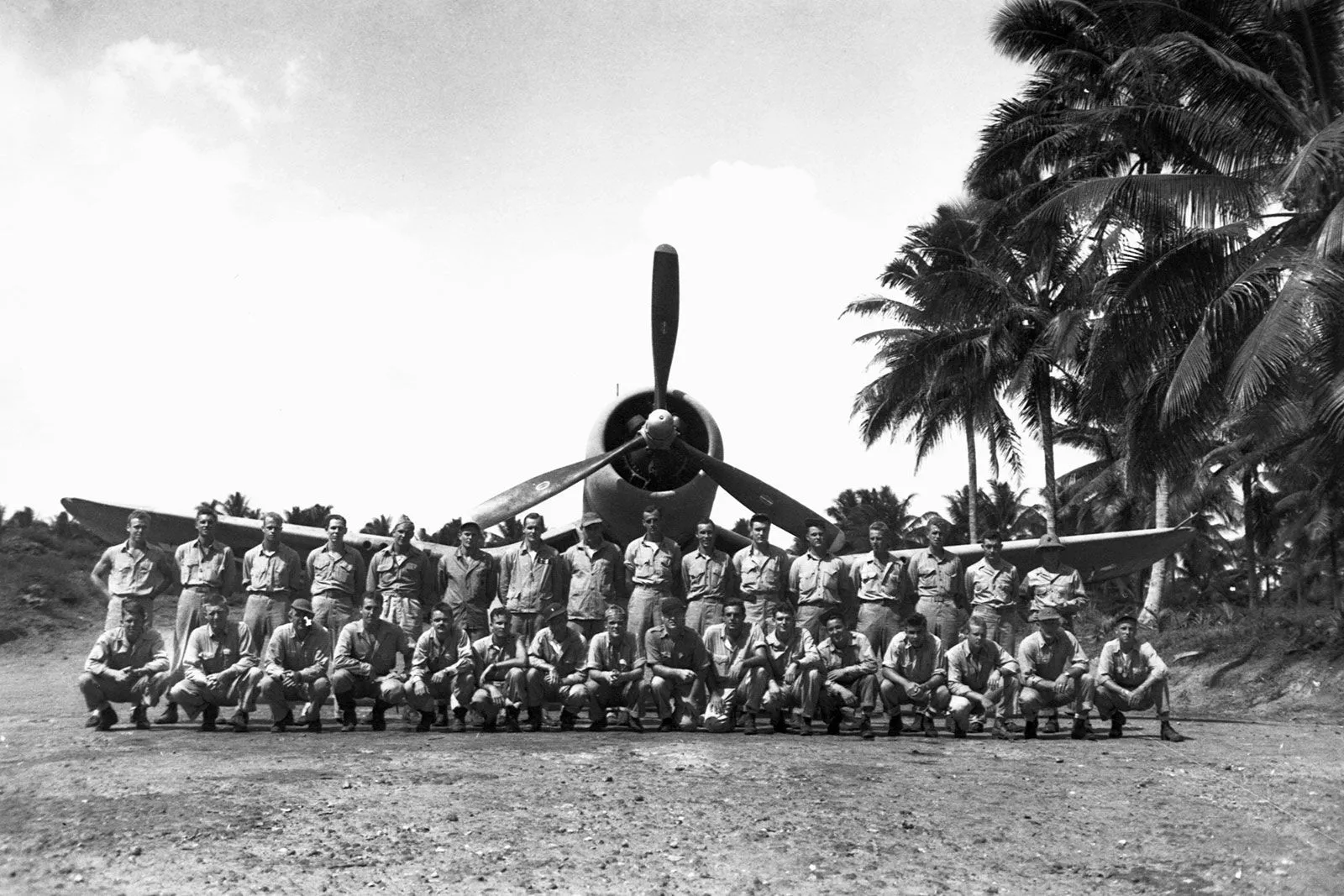 طاقم طائرة F4U Corsair الأمريكية يقفون أمام الطائرة في موقع استوائي خلال الحرب العالمية الثانية، مع أشجار نخيل في الخلفية.
