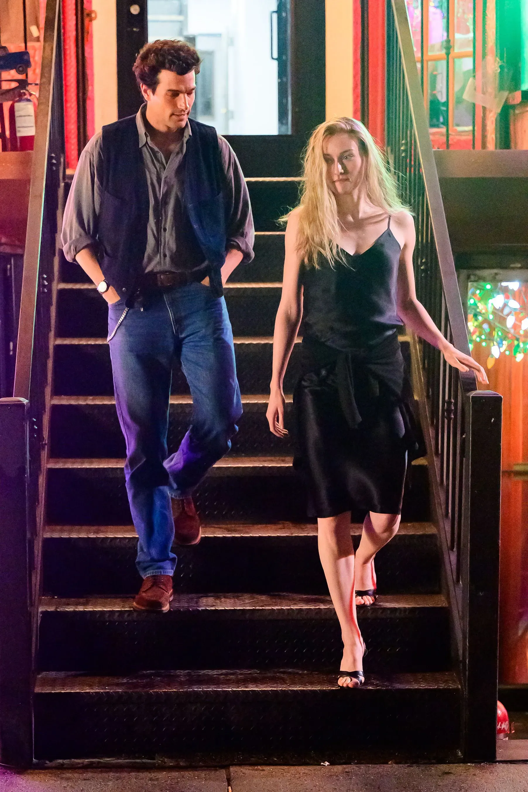 امرأة ترتدي فستانًا أسود بسيطًا تسير على الدرج مع رجل يرتدي سترة، في مشهد يعكس أناقة غير متكلفة.