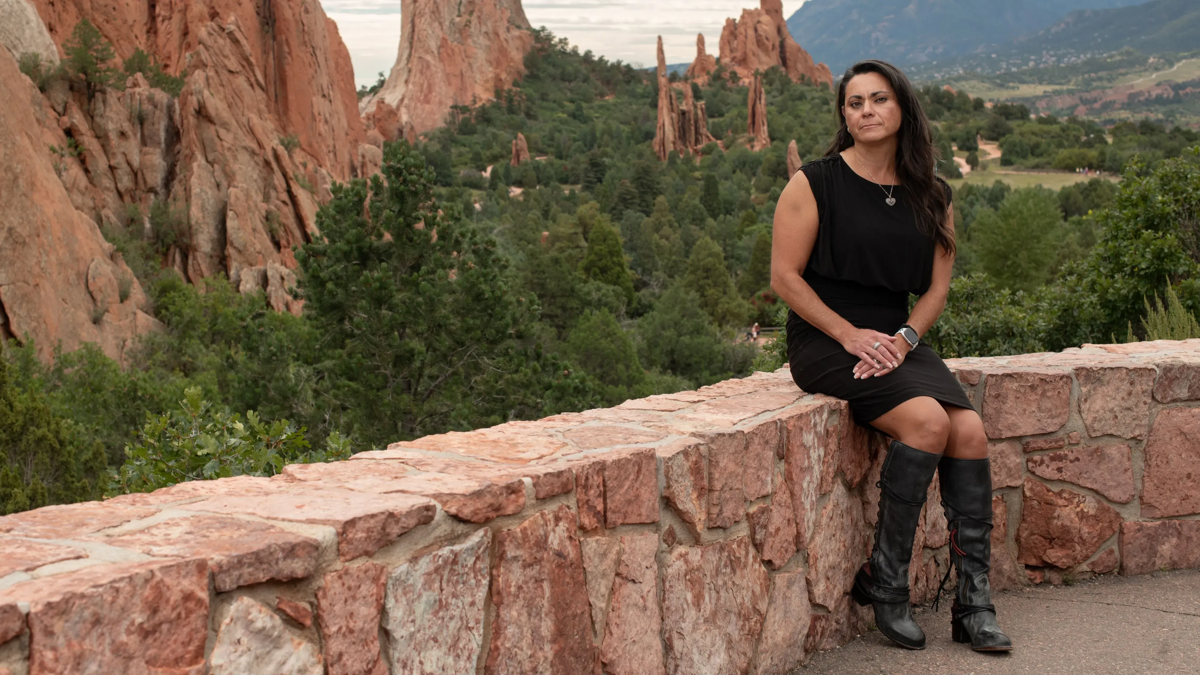 امرأة تجلس على حافة حجرية في منطقة طبيعية، محاطة بتضاريس صخرية وأشجار، تعبر عن تأمل أو تفكير عميق في خلفية الجبال.