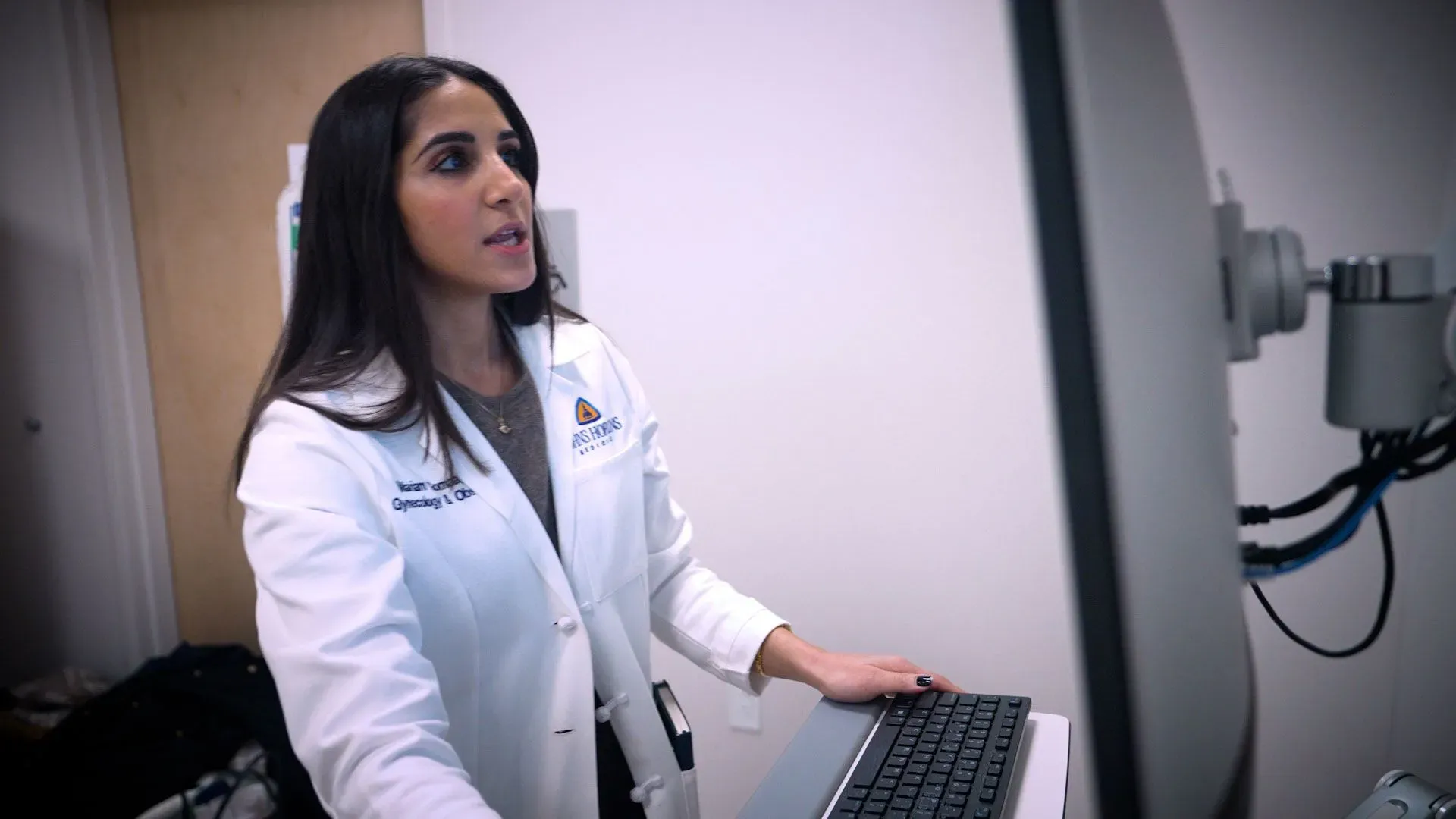 طبيبة تتحدث في عيادة، ترتدي معطفًا أبيض وتستخدم جهاز كمبيوتر، مما يعكس النقاش حول وسائل منع الحمل الهرمونية.