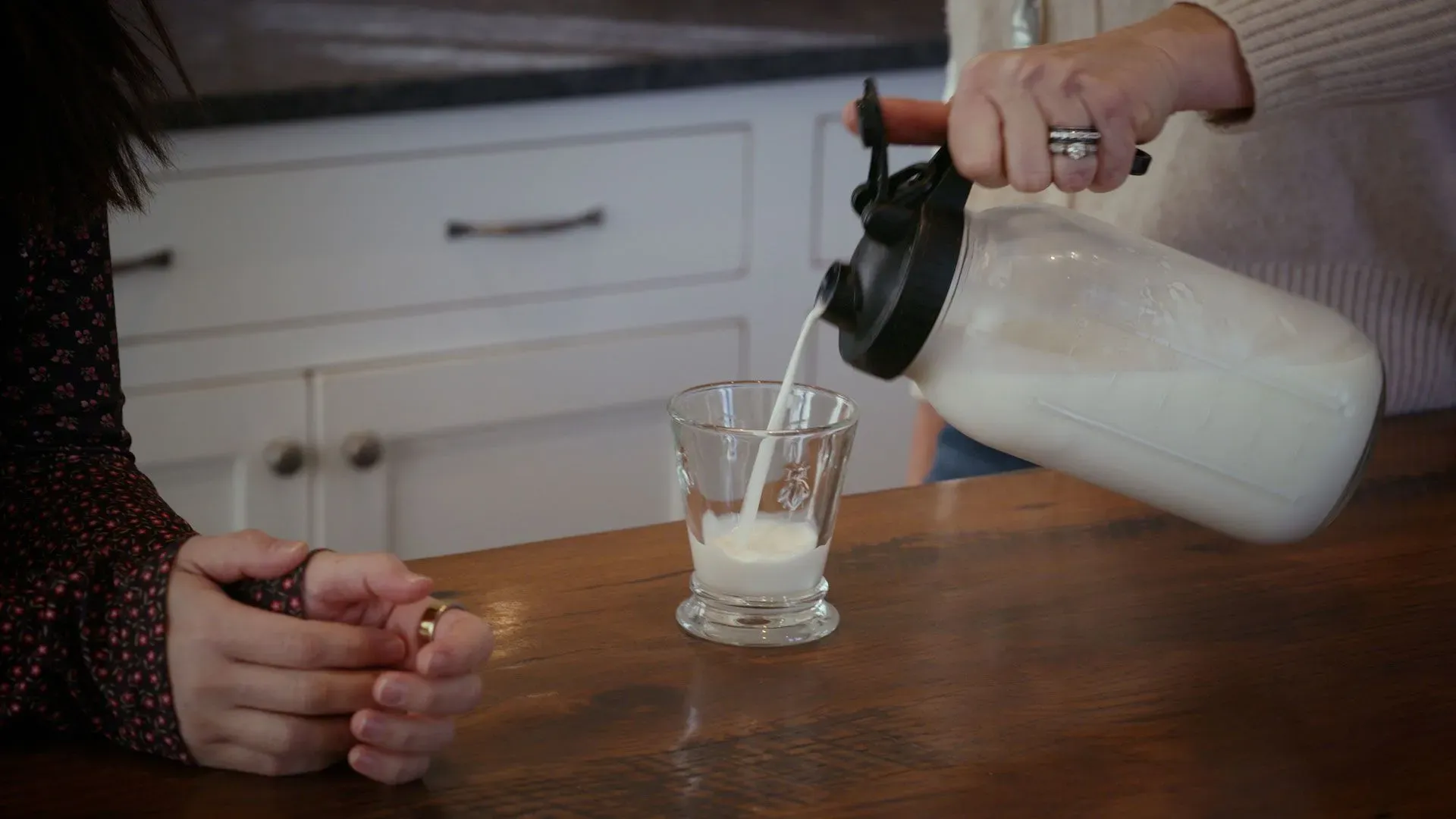 امرأة تصب الحليب في كوب زجاجي، بينما تجلس امرأة أخرى على الطاولة، مما يعكس لحظة من التفاعل اليومي.