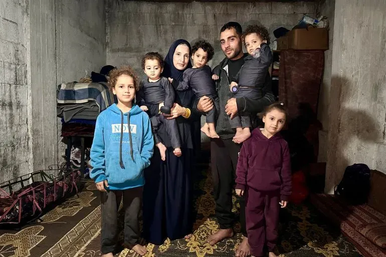 عائلة مكونة من سبعة أفراد، تتكون من والدين وخمسة أطفال، في غرفة بسيطة في دير البلح، تعكس تحديات الحياة اليومية بعد الحرب.