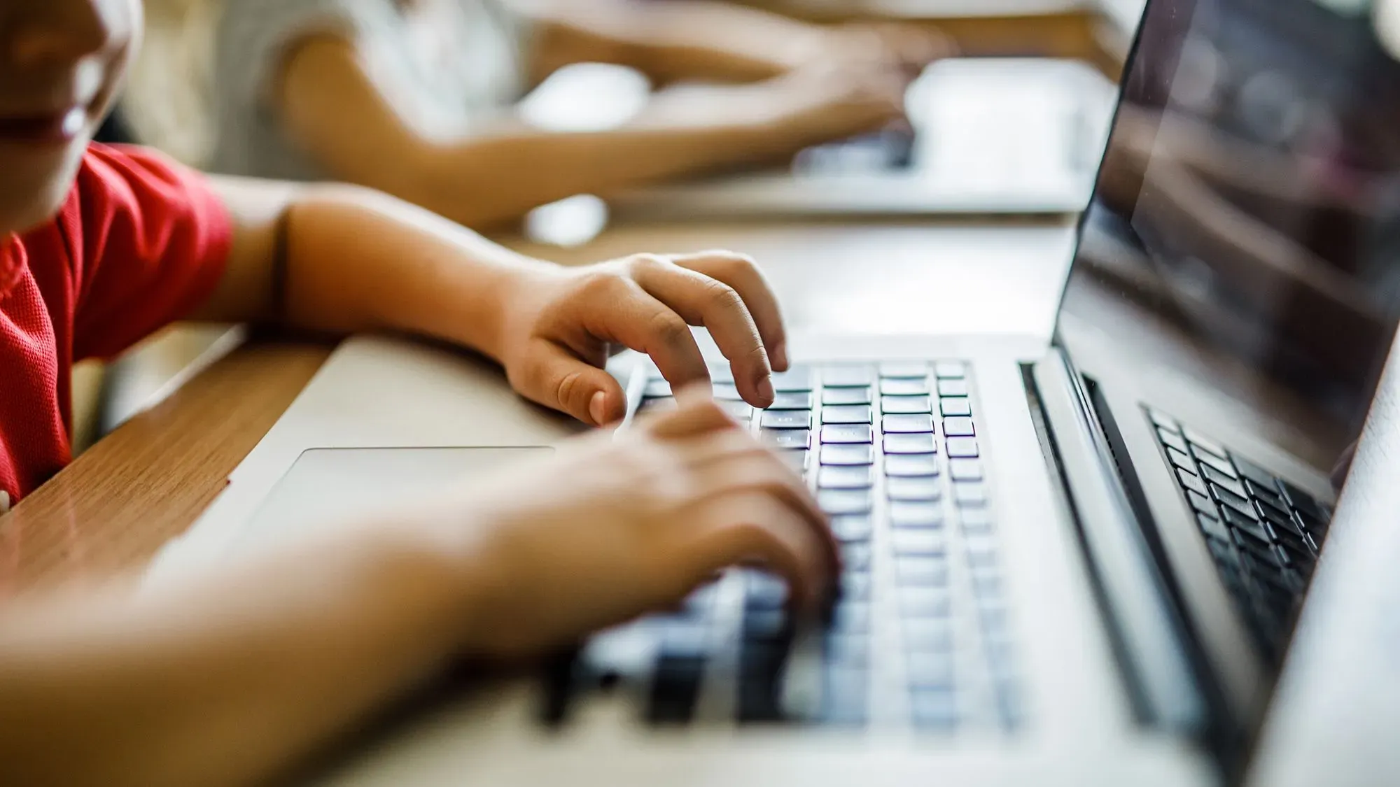 طلاب يستخدمون أجهزة الكمبيوتر المحمولة في فصل دراسي، مع التركيز على الأيدي التي تكتب على لوحة المفاتيح، مما يعكس دمج الذكاء الاصطناعي في التعليم.