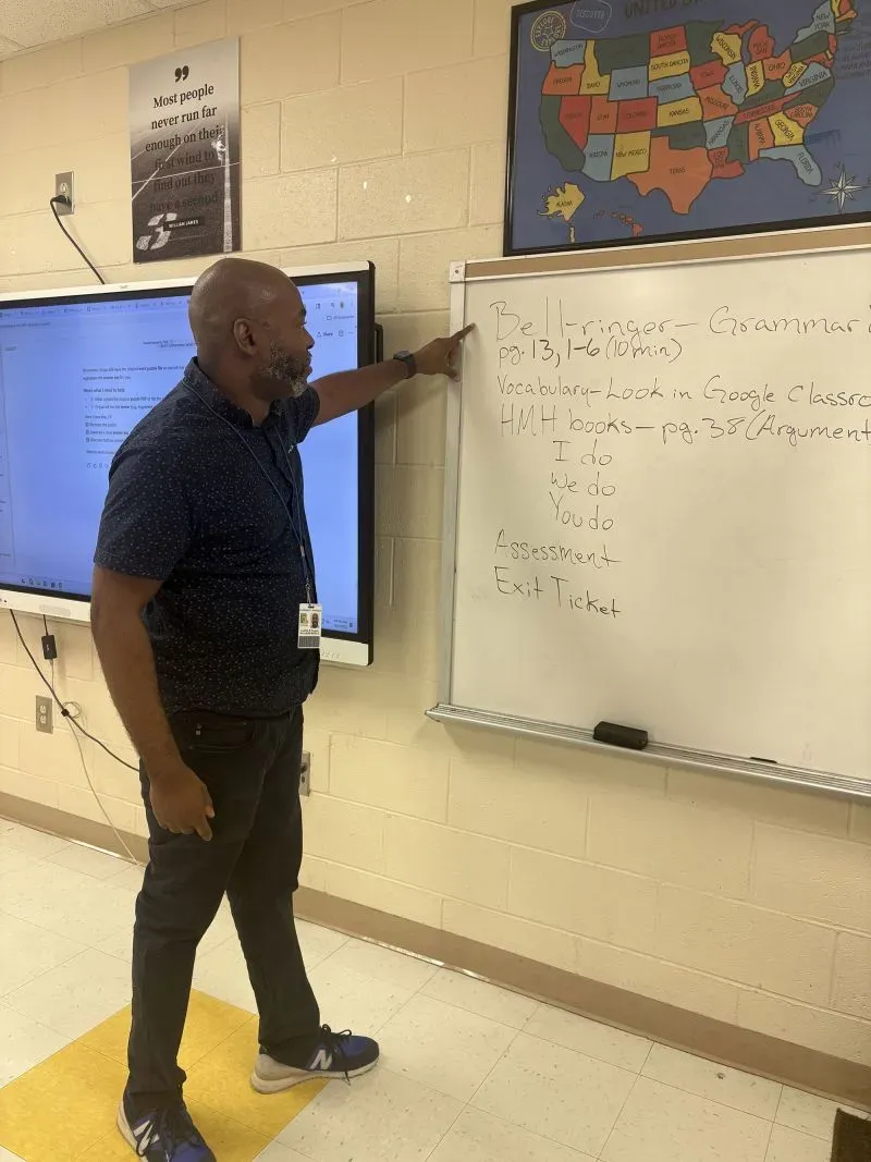 مدرس يشرح درسًا في فصل دراسي، مع وجود لوحة بيضاء تحتوي على معلومات عن الأنشطة والتقييمات، تعكس استخدام الذكاء الاصطناعي في التعليم.