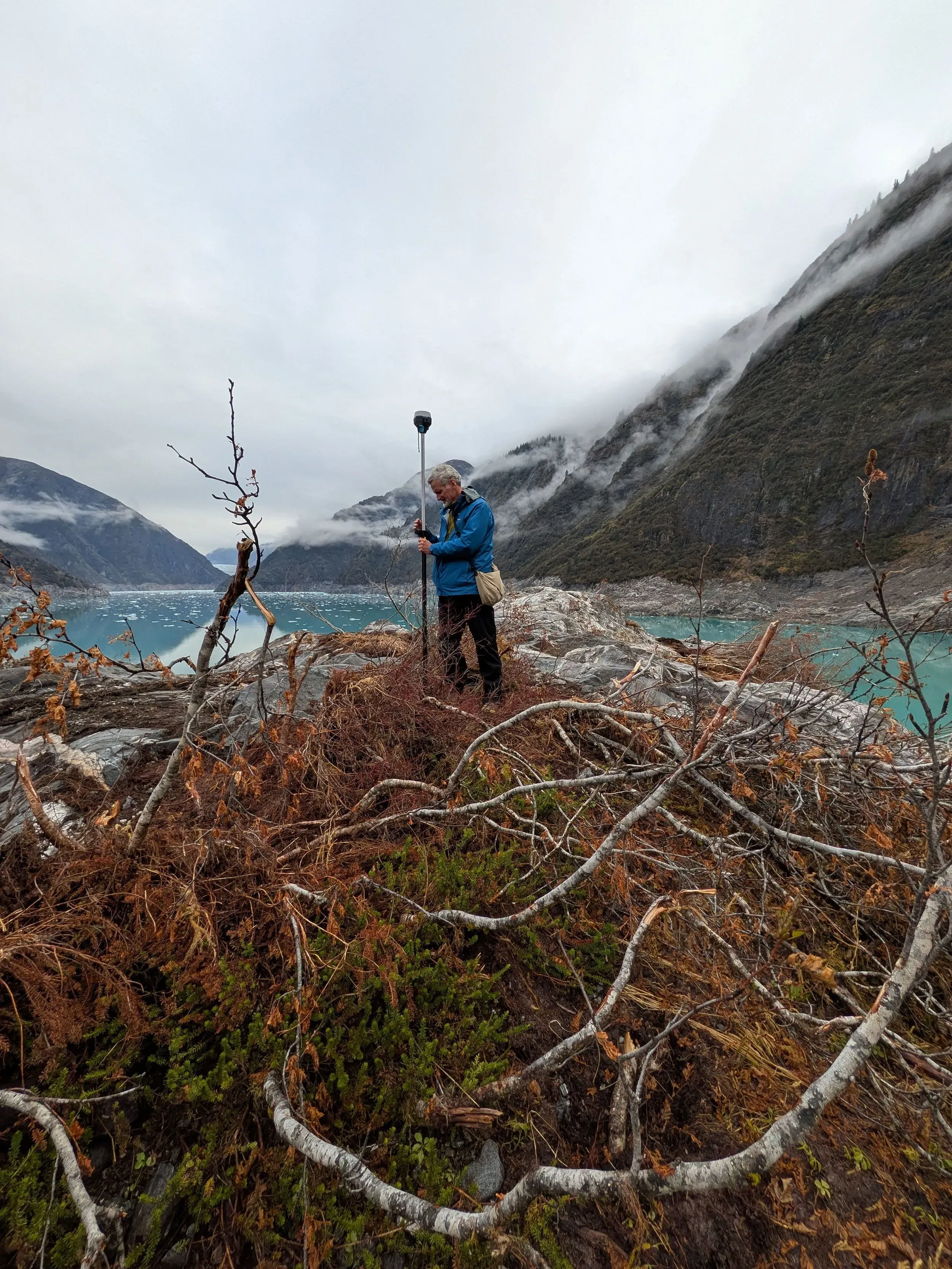 عالم زلازل يقف على صخور عارية قرب مضيق تريسي آرم في ألاسكا، محاطًا بأشجار ميتة، بعد انهيار أرضي هائل.