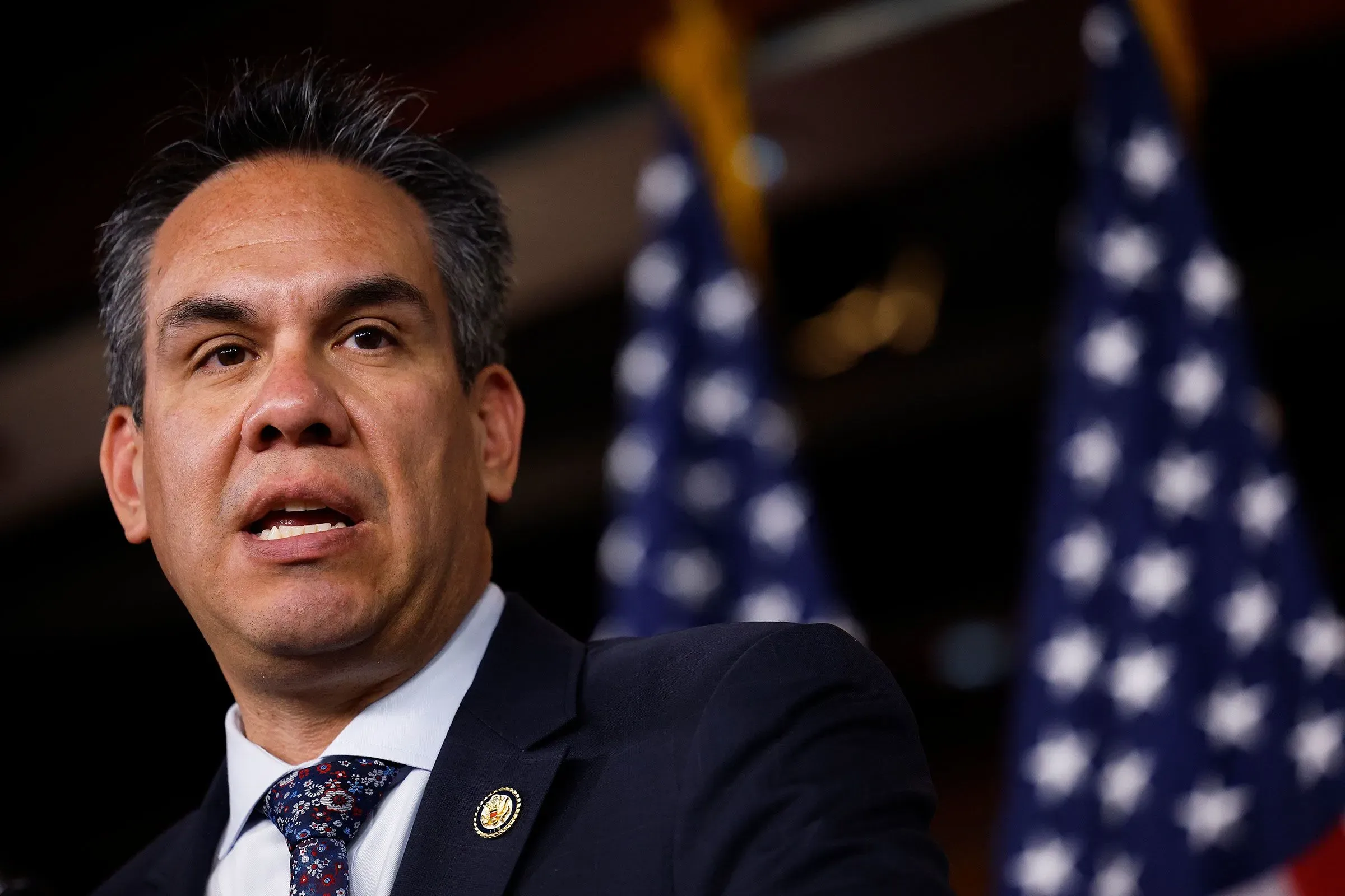 نائب ديمقراطي يتحدث خلال مؤتمر صحفي، مع وجود أعلام أمريكية خلفه، في سياق مناقشة أحداث 6 يناير 2021 وتأثيرها السياسي.