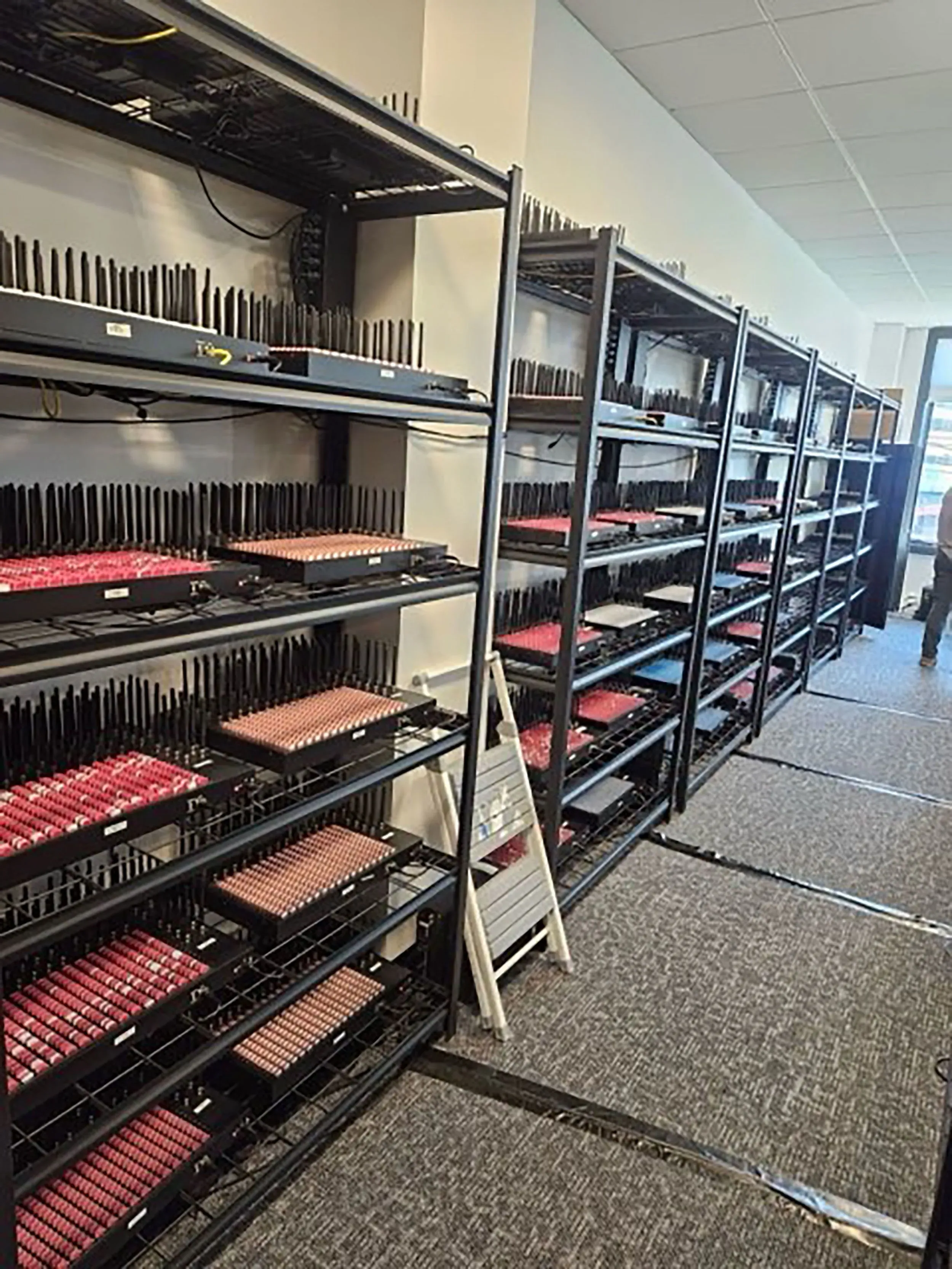 صورة لمستودع يحتوي على صفوف من أجهزة الاتصال، بما في ذلك شريحة هاتف محمول، تُظهر شبكة معقدة من الأجهزة المستخدمة في عمليات سواتينغ.